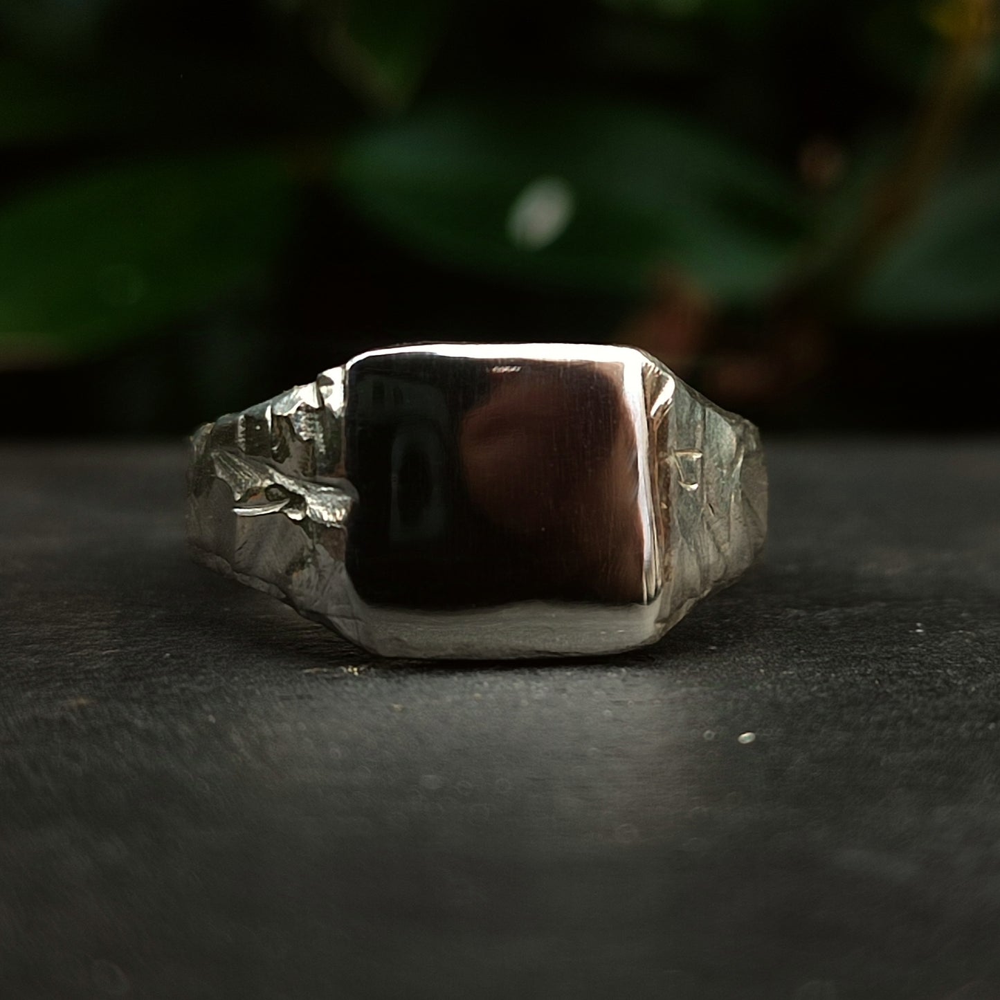 The Square Rock Signet by silverhollowjewellery, crafted from recycled sterling silver with a plain rectangular face, is displayed on a dark surface with a blurred green background.