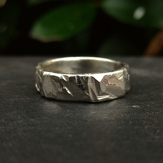 The Thick Rock Band by silverhollowjewellery, crafted from recycled sterling silver with a hammered texture, is displayed on a dark surface with blurred green leaves in the background.