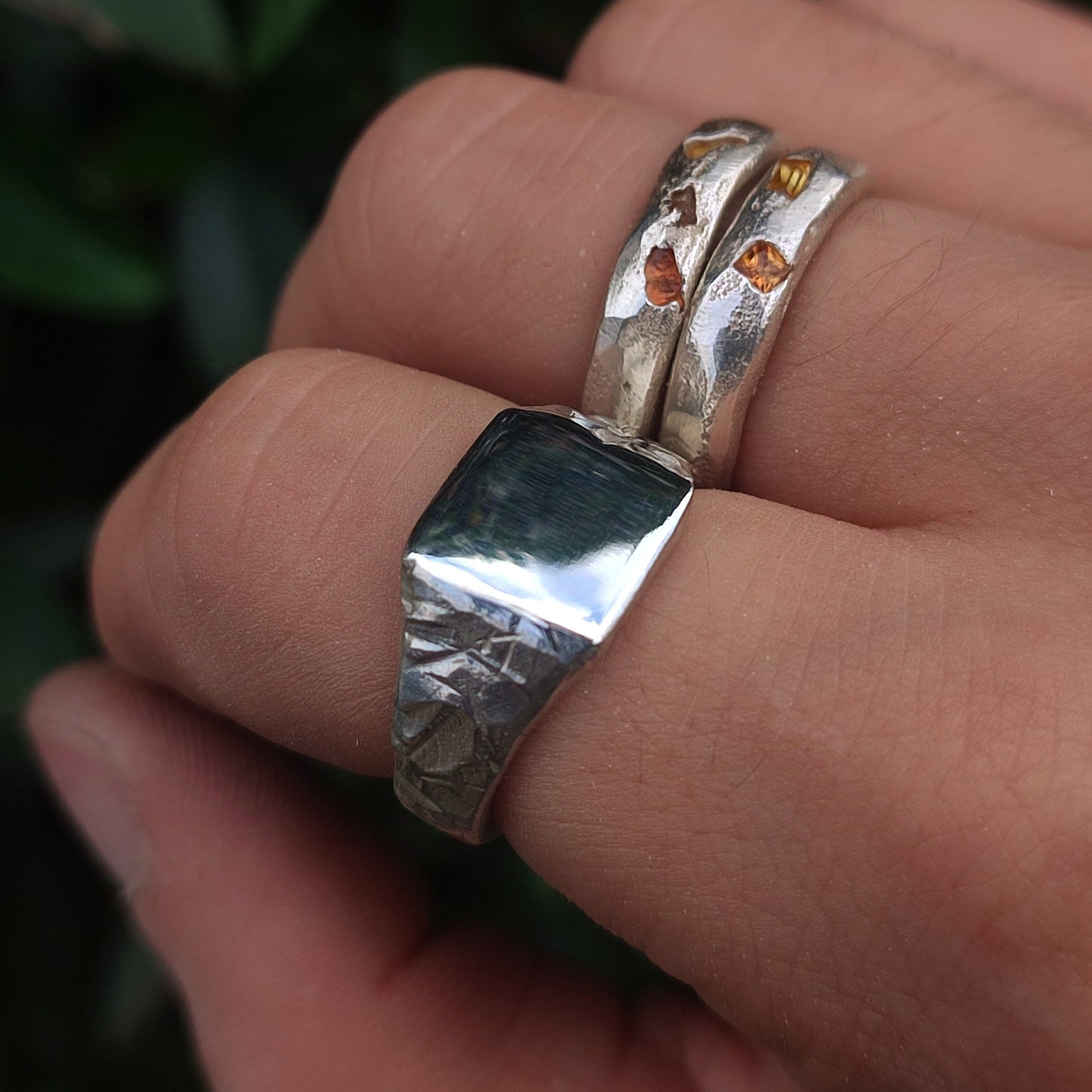 A hand wears two recycled sterling silver rings from silverhollowjewellery: the Square Rock Signet, a smooth, rectangular piece, and another with amber-colored stones and a subtle rock-textured finish.