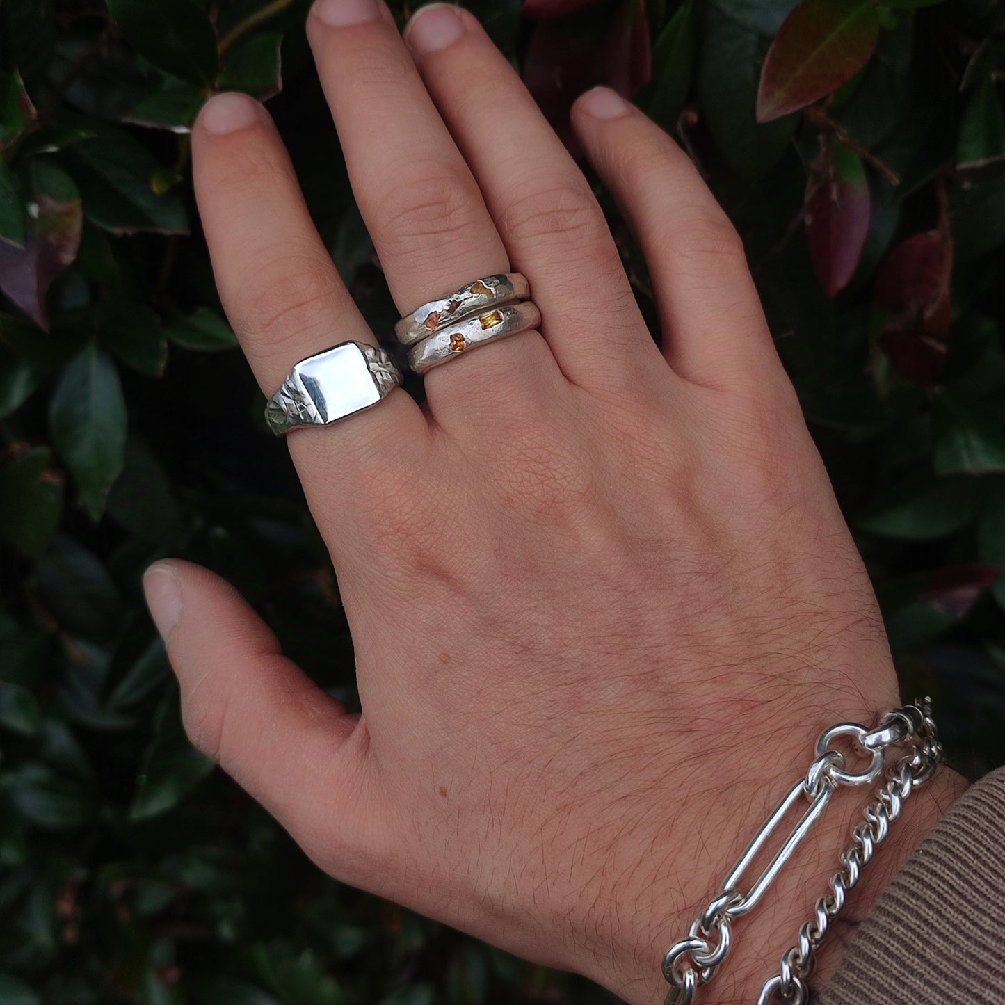 A hand wearing three silver rings—featuring the Square Rock Signet by silverhollowjewellery—and a silver chain bracelet is held against a leafy green background.