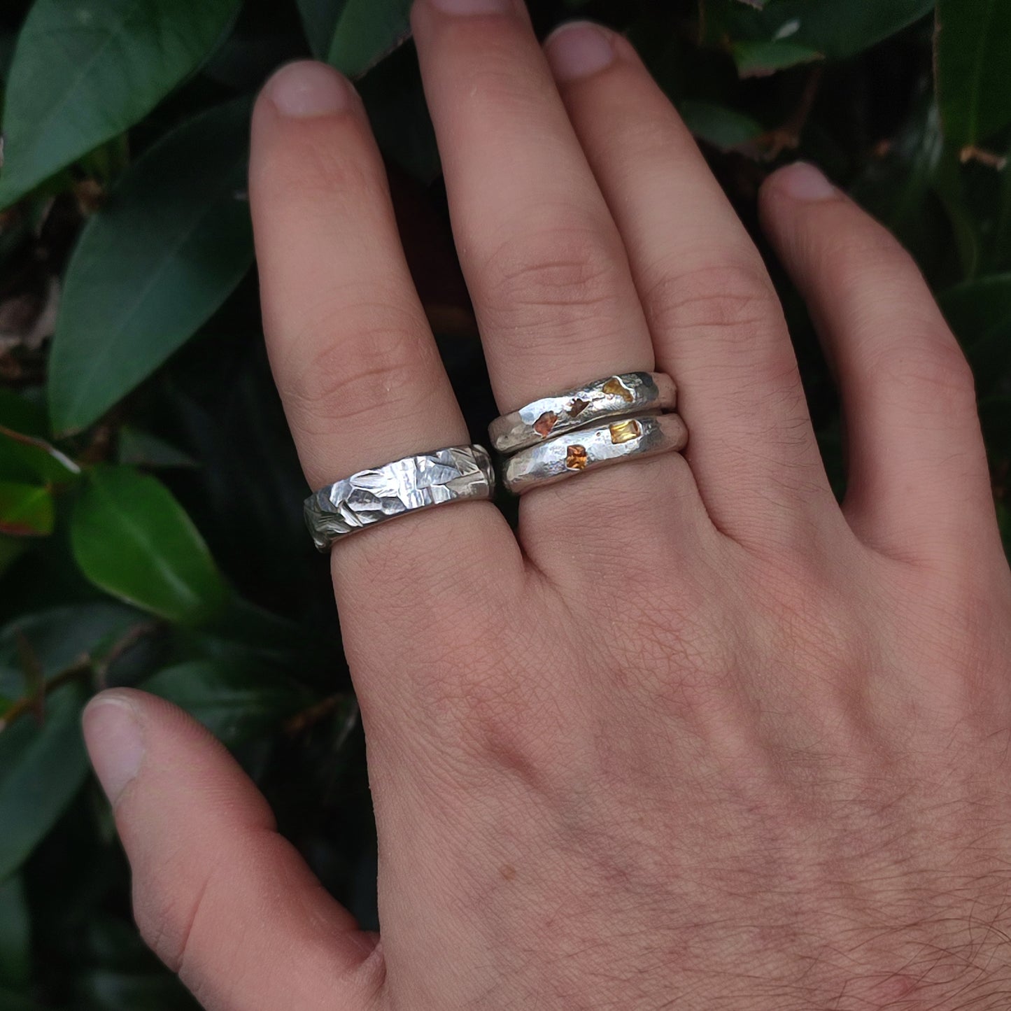 A hand wears three recycled 925 sterling silver rings from silverhollowjewellery, including the Thick Rock Band—a chunky ring with rock-textured details and gold accents—set against green leaves.