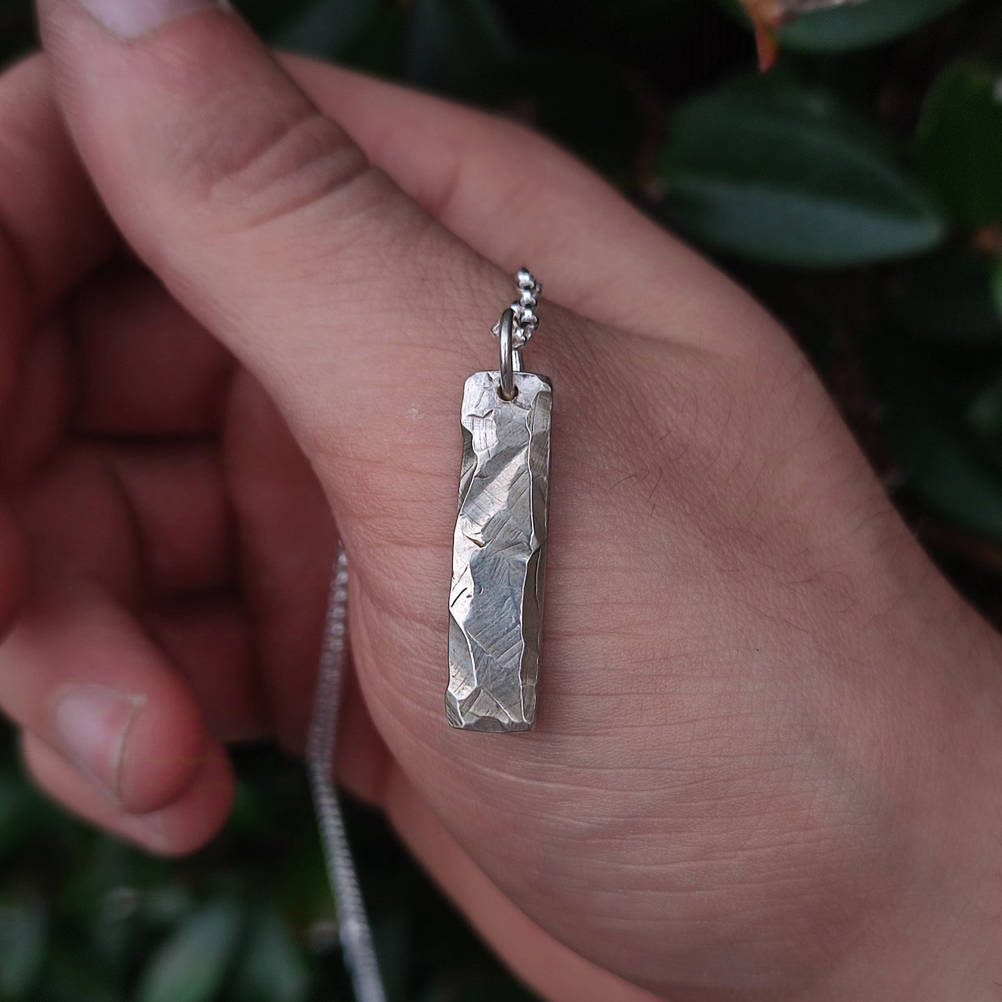 A hand holds the silverhollowjewellery Rock Pendant—a rectangular, rock-textured piece crafted from recycled sterling silver—hanging on a chain, with green leaves in the background.
