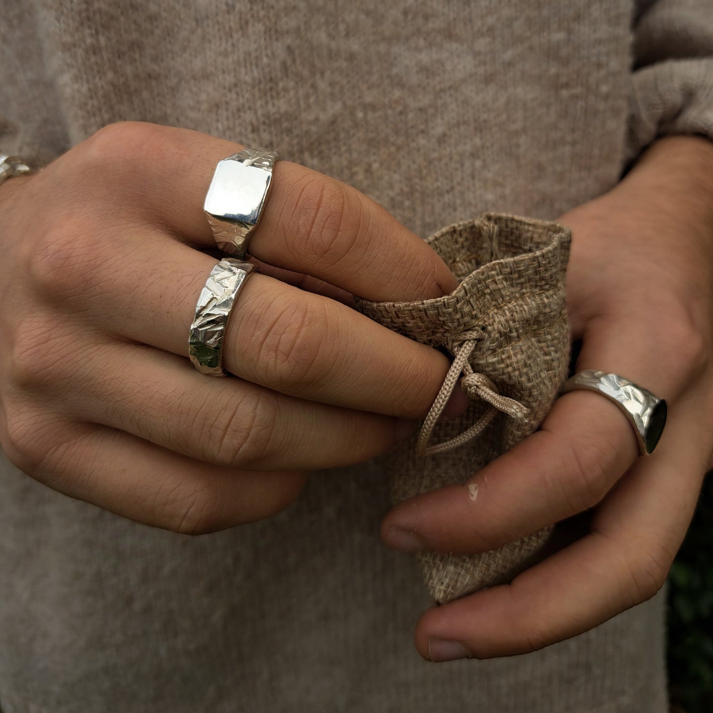 A person wearing multiple sterling silver rings, including the Thick Rock Band by silverhollowjewellery, holds a small textured drawstring pouch.