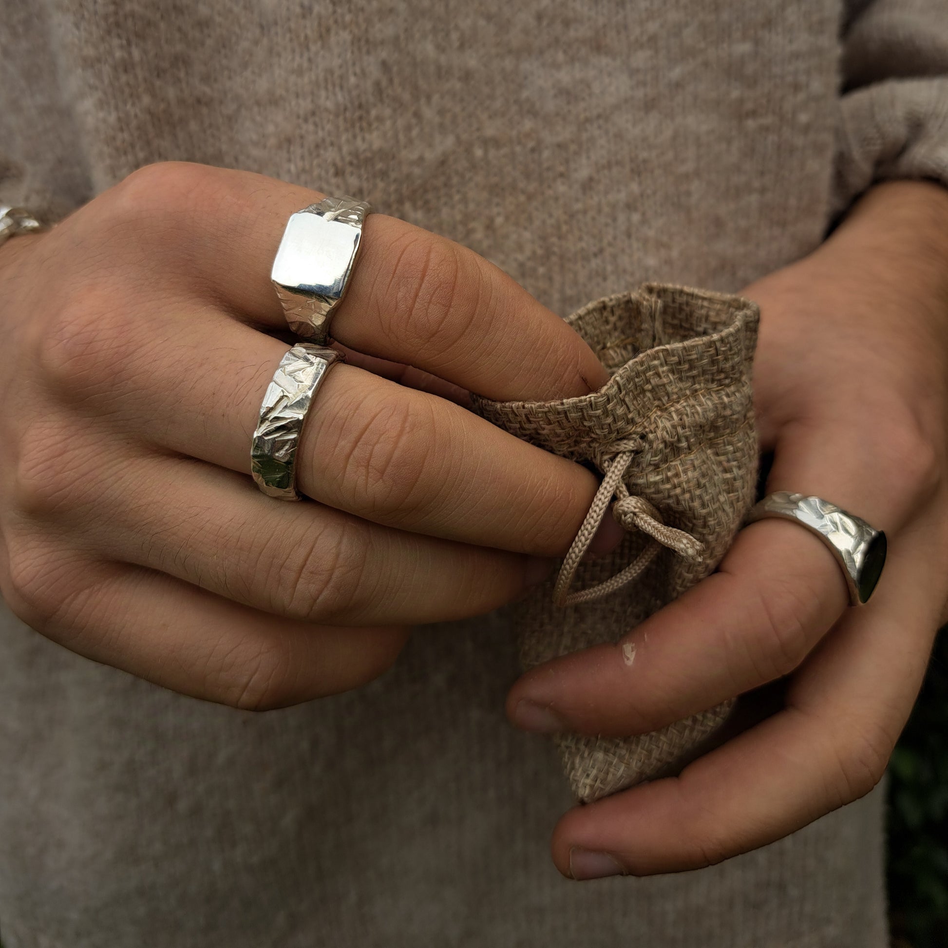 A person wearing three silver rings, including the silverhollowjewellery Square Rock Signet, holds a small burlap drawstring pouch.