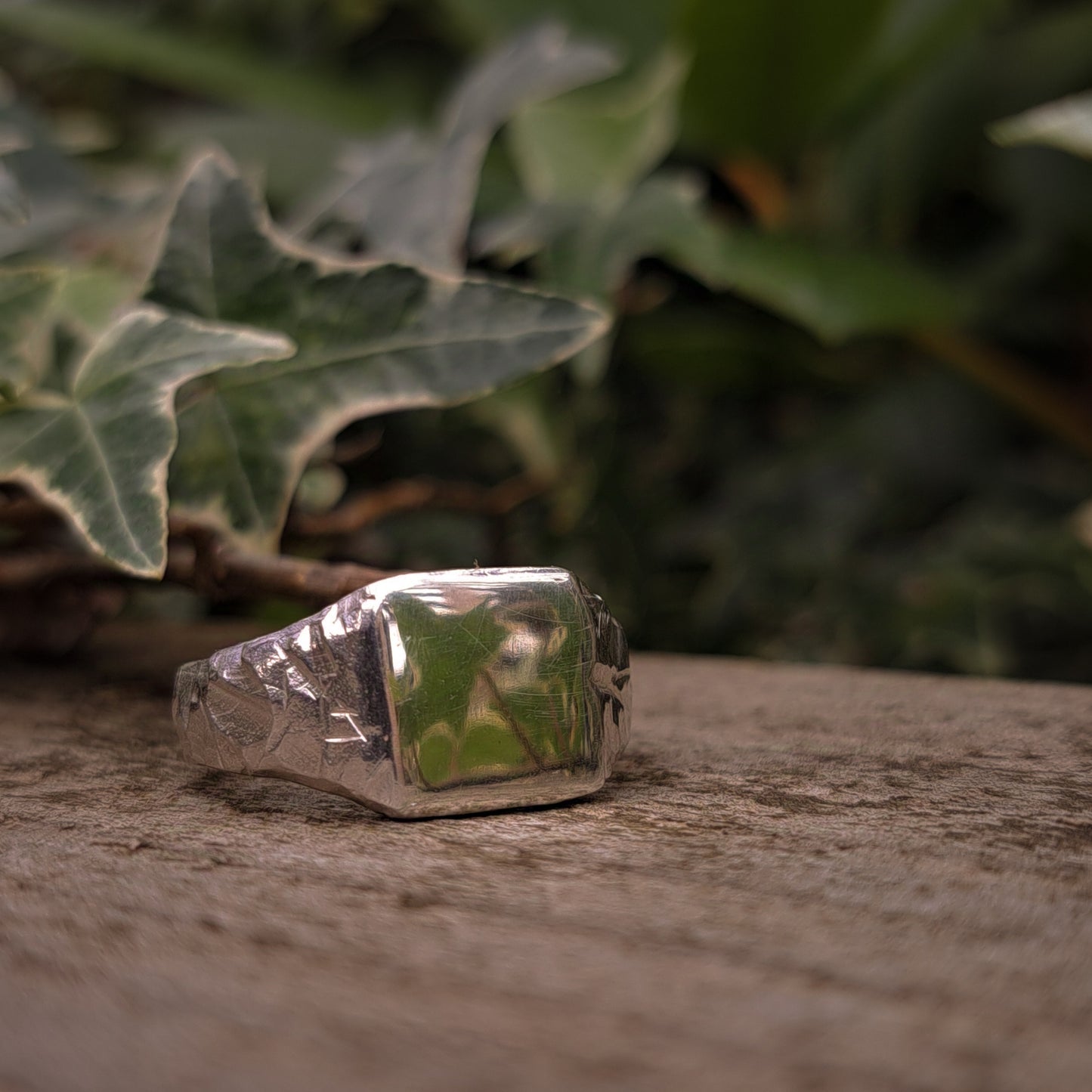 The Square Rock Signet by silverhollowjewellery, made from recycled sterling silver and featuring an engraved design, rests on a wooden surface with green ivy leaves in the background.