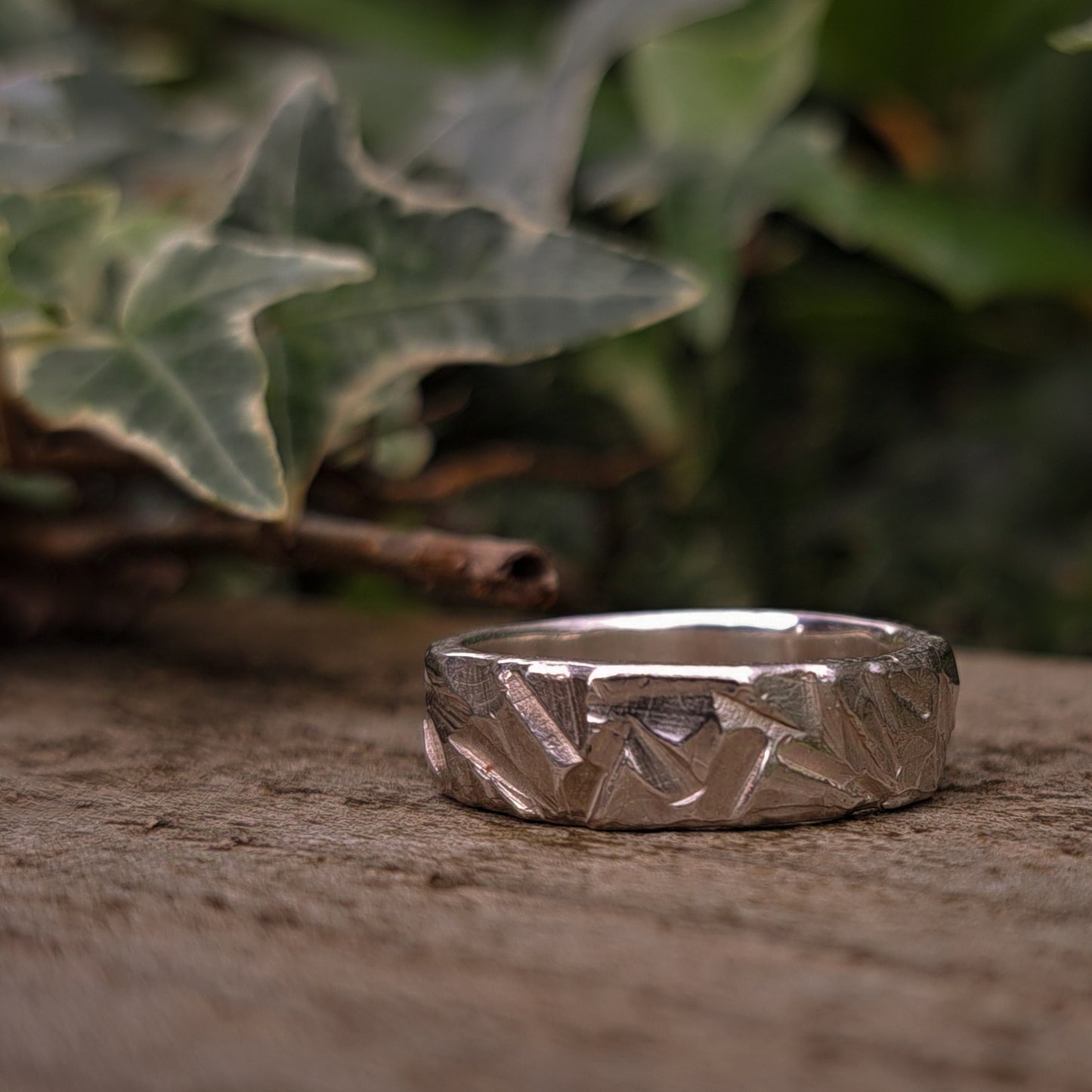 The Thick Rock Band by silverhollowjewellery, a sterling silver ring with a textured, rock-inspired design, sits on a wooden surface with blurred green ivy leaves in the background.