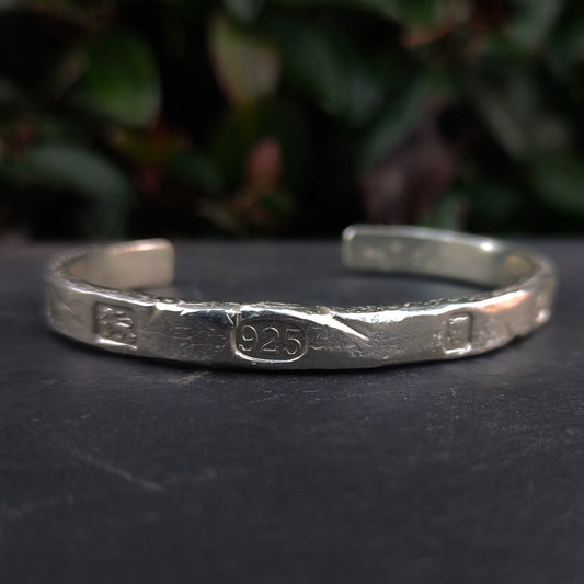The Molten Rock Cuff by silverhollowjewellery is a distressed, hammered bracelet made from recycled sterling silver with "925" stamped at the center. Blurred green foliage appears in the background.