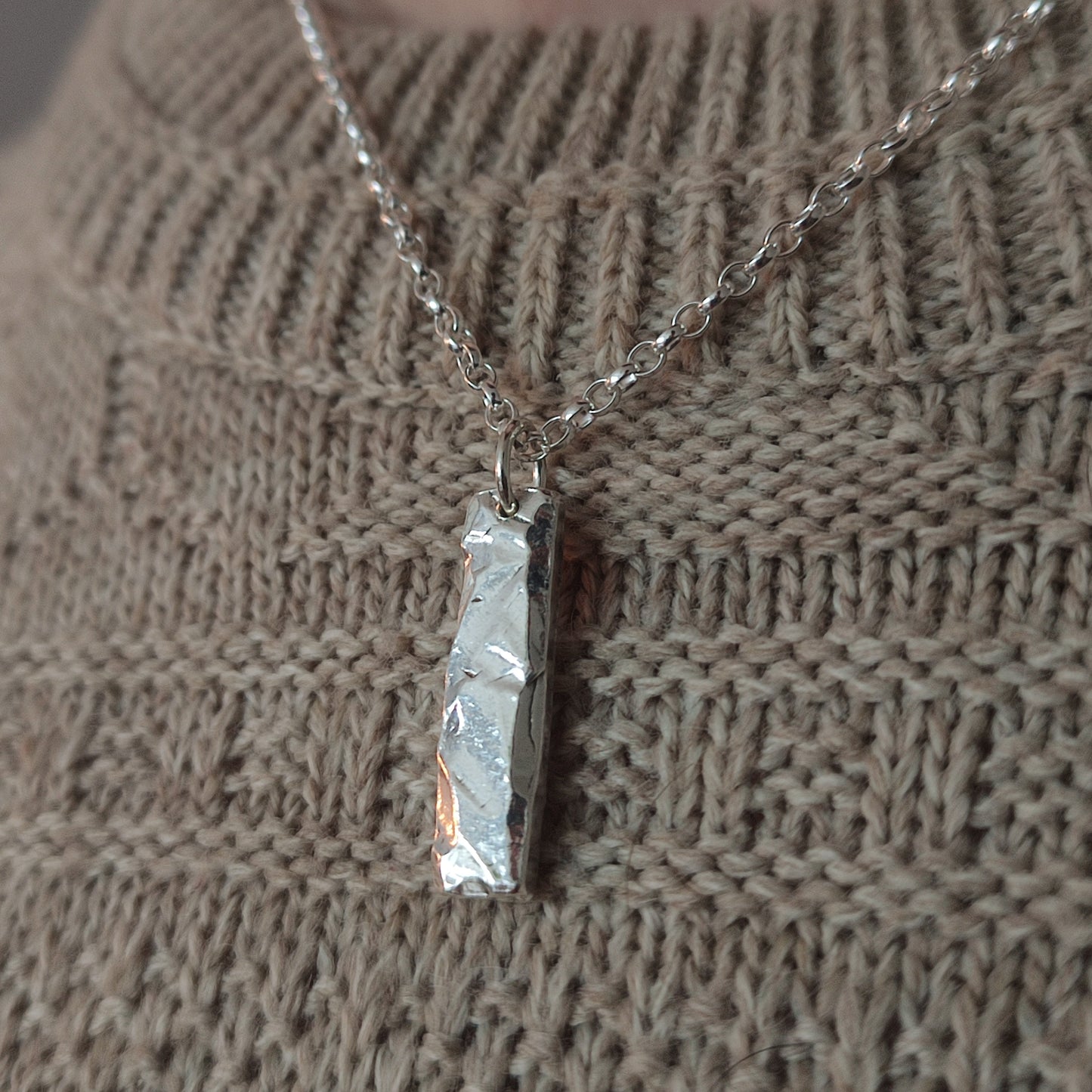 The Rock Pendant by silverhollowjewellery, featuring a rock-textured silver surface, hangs on a chain over a beige knitted sweater.