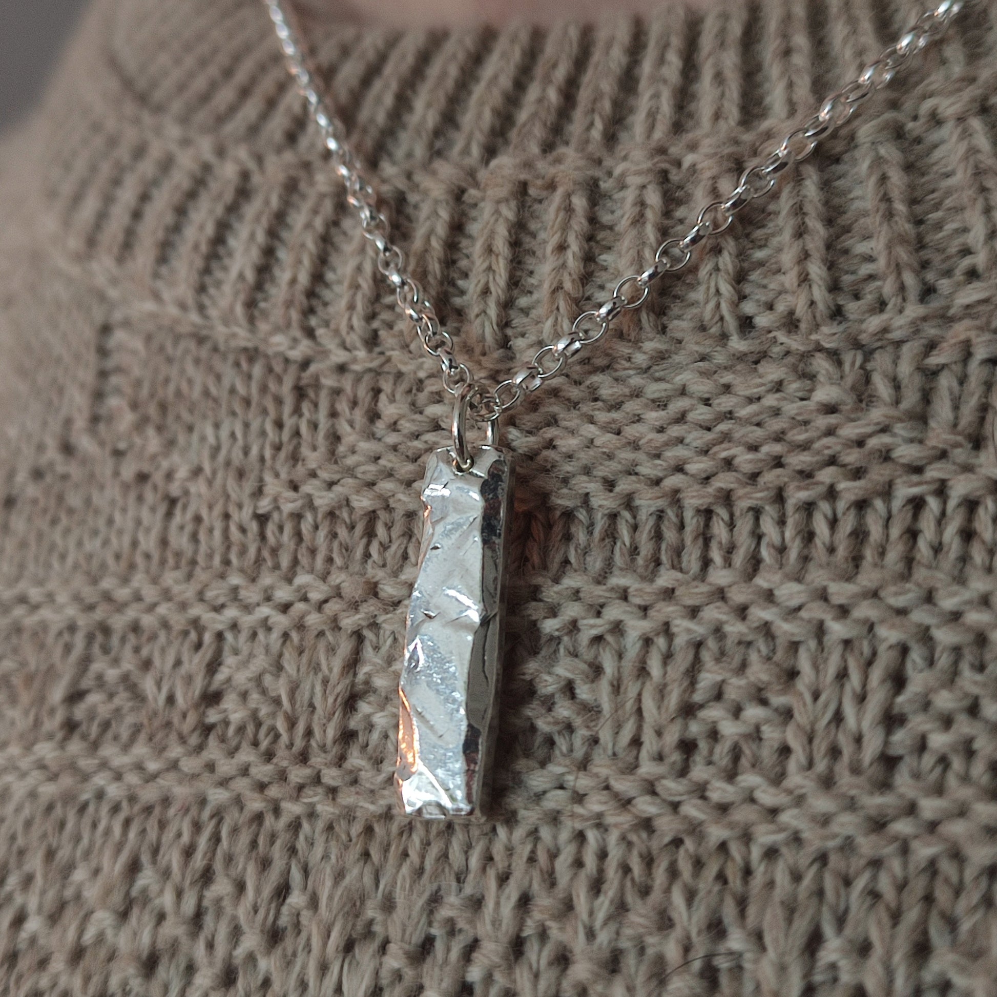 The Rock Pendant by silverhollowjewellery, featuring a rock-textured silver surface, hangs on a chain over a beige knitted sweater.