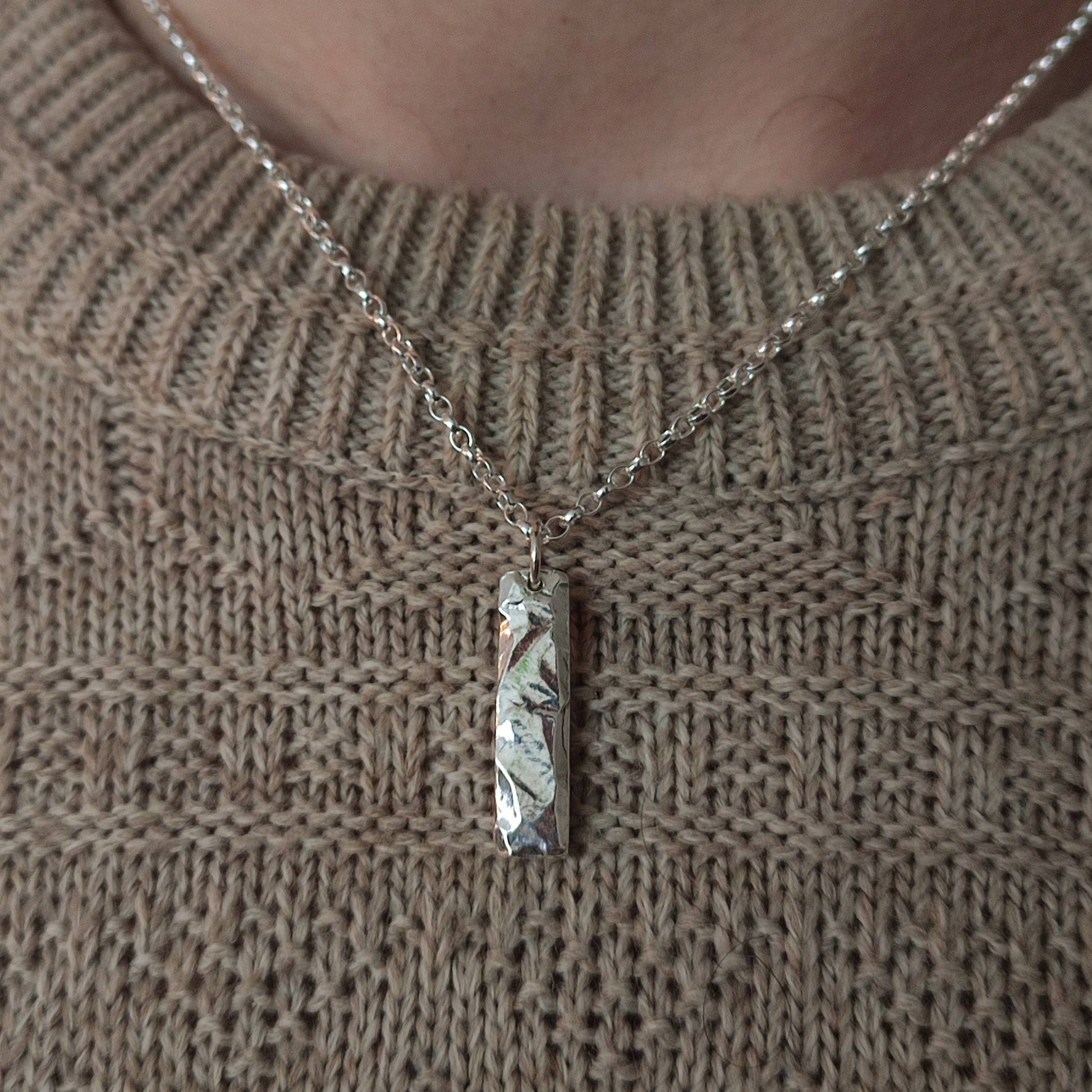 Someone wears the silverhollowjewellery Rock Pendant, a solid sterling silver necklace with a rectangular rock-textured pendant, over a beige knitted sweater.