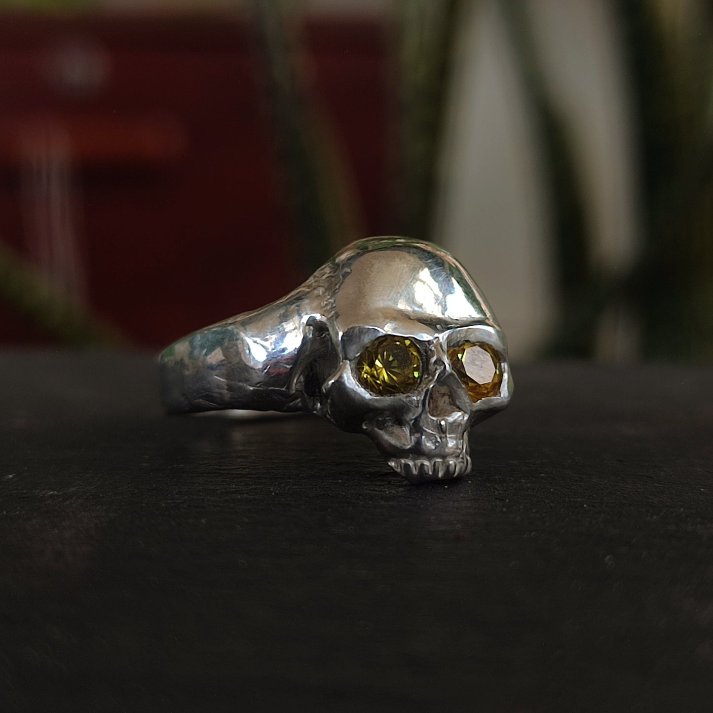 The Yellow Sapphire Skull Ring by silverhollowjewellery is crafted from recycled sterling silver, featuring two yellow sapphires as eyes and shown on a dark surface with plants in the background.