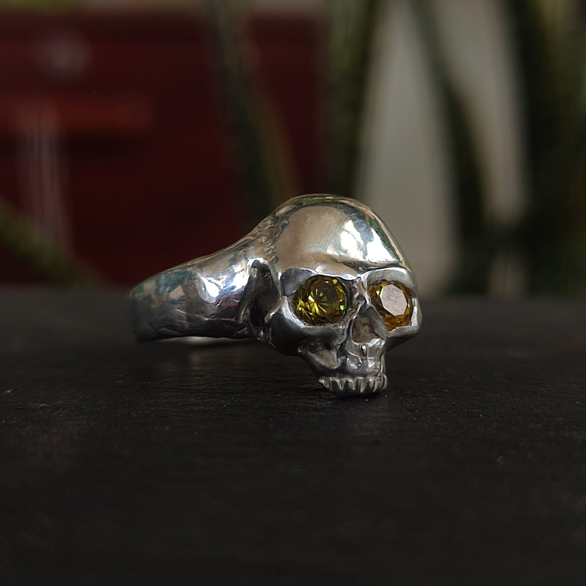 The Yellow Sapphire Skull Ring by silverhollowjewellery is crafted from recycled sterling silver, featuring two yellow sapphires as eyes and shown on a dark surface with plants in the background.