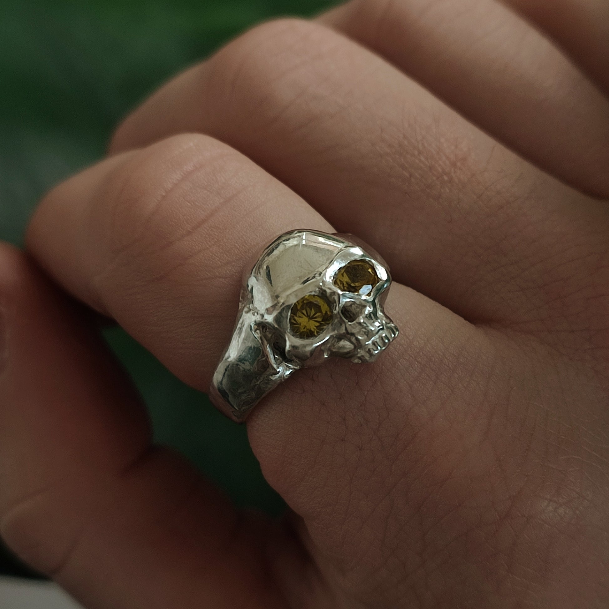 A hand displays the silverhollowjewellery Yellow Sapphire Skull Ring, crafted from recycled sterling silver with yellow sapphire eyes, set against a blurred green background.