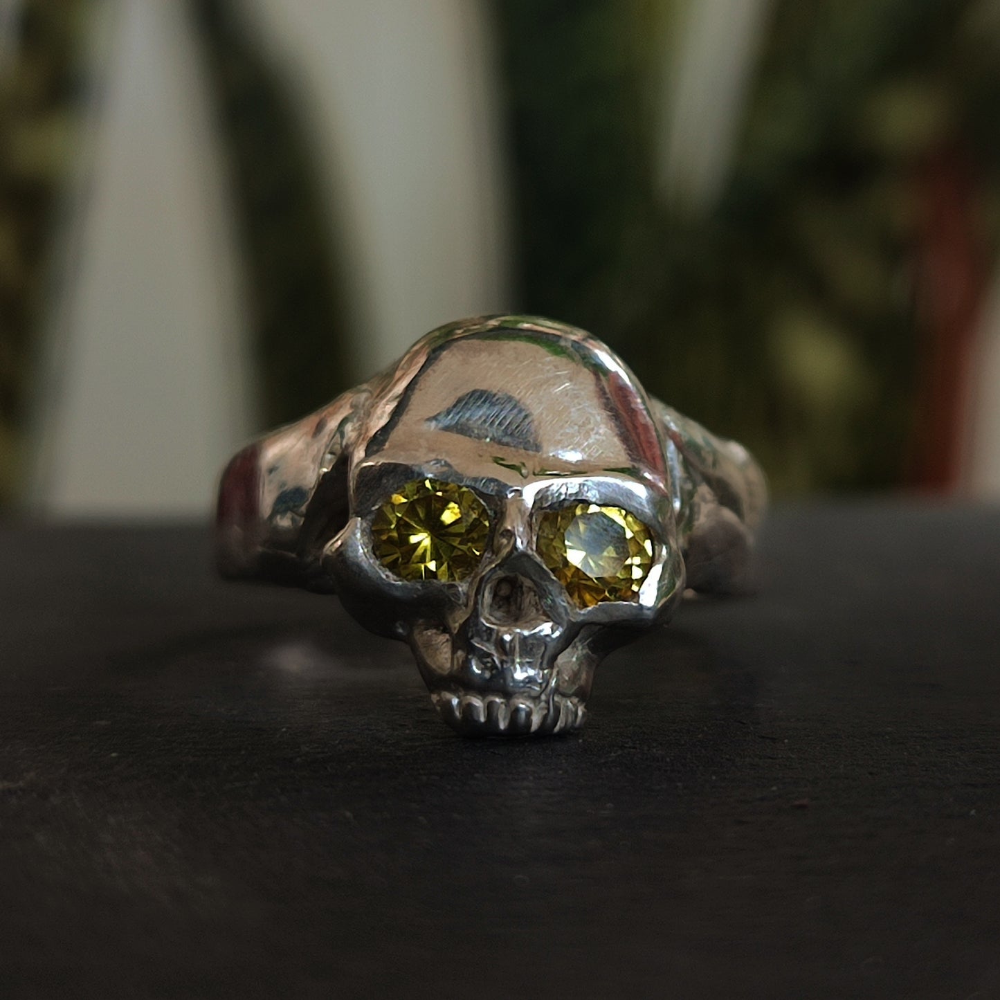 The Yellow Sapphire Skull Ring by silverhollowjewellery, made from recycled sterling silver and featuring yellow sapphire eyes, is displayed on a dark surface with blurred green plants in the background.