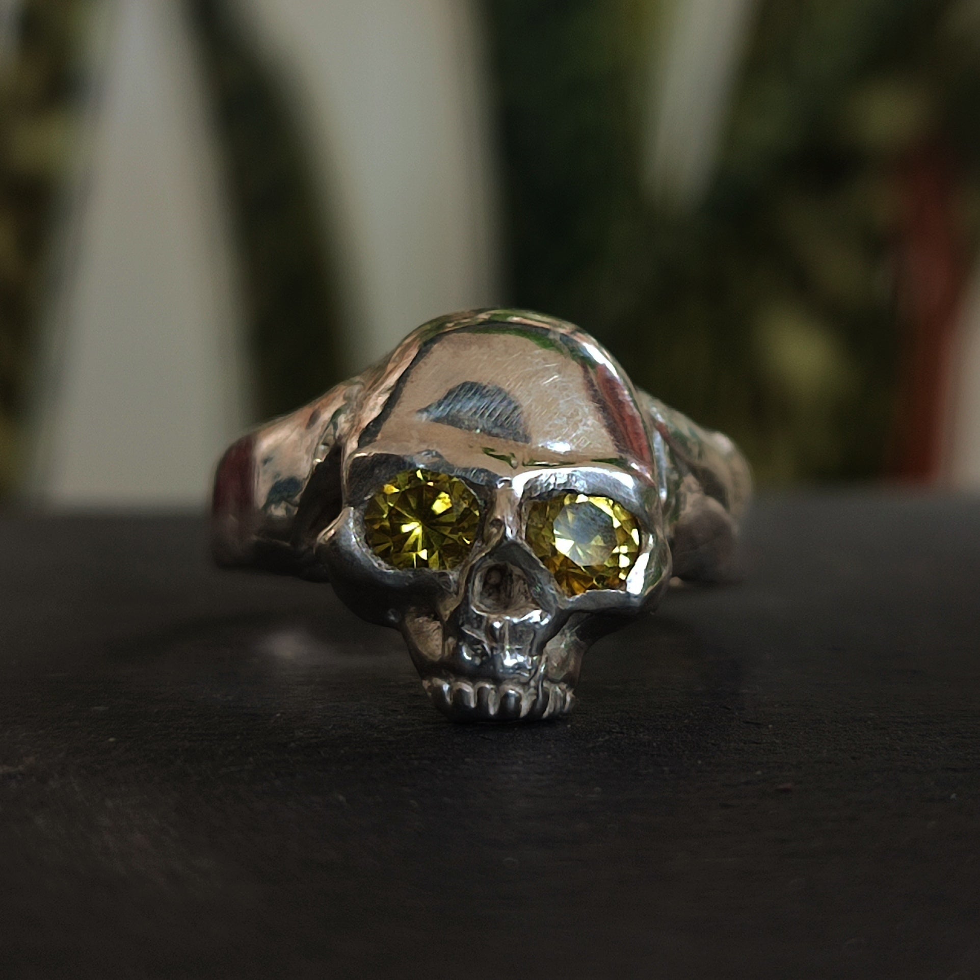 The Yellow Sapphire Skull Ring by silverhollowjewellery, made from recycled sterling silver and featuring yellow sapphire eyes, is displayed on a dark surface with blurred green plants in the background.