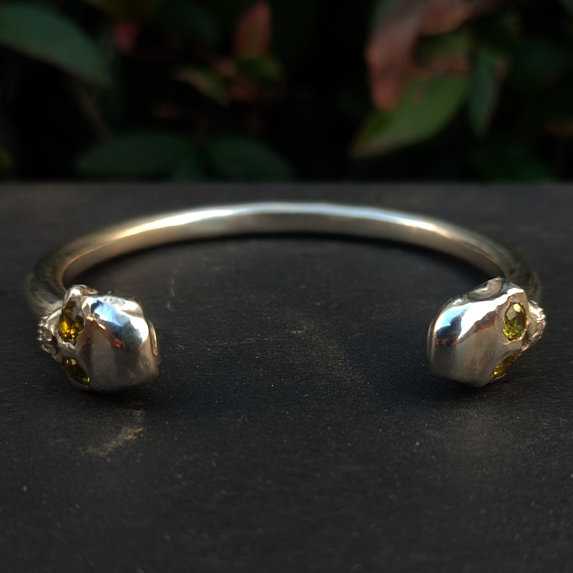 The Yellow Sapphire Skull Cuff by silverhollowjewellery features two skull-shaped ends with yellow sapphire eyes, crafted from recycled sterling silver and shown against a dark background with blurred foliage.