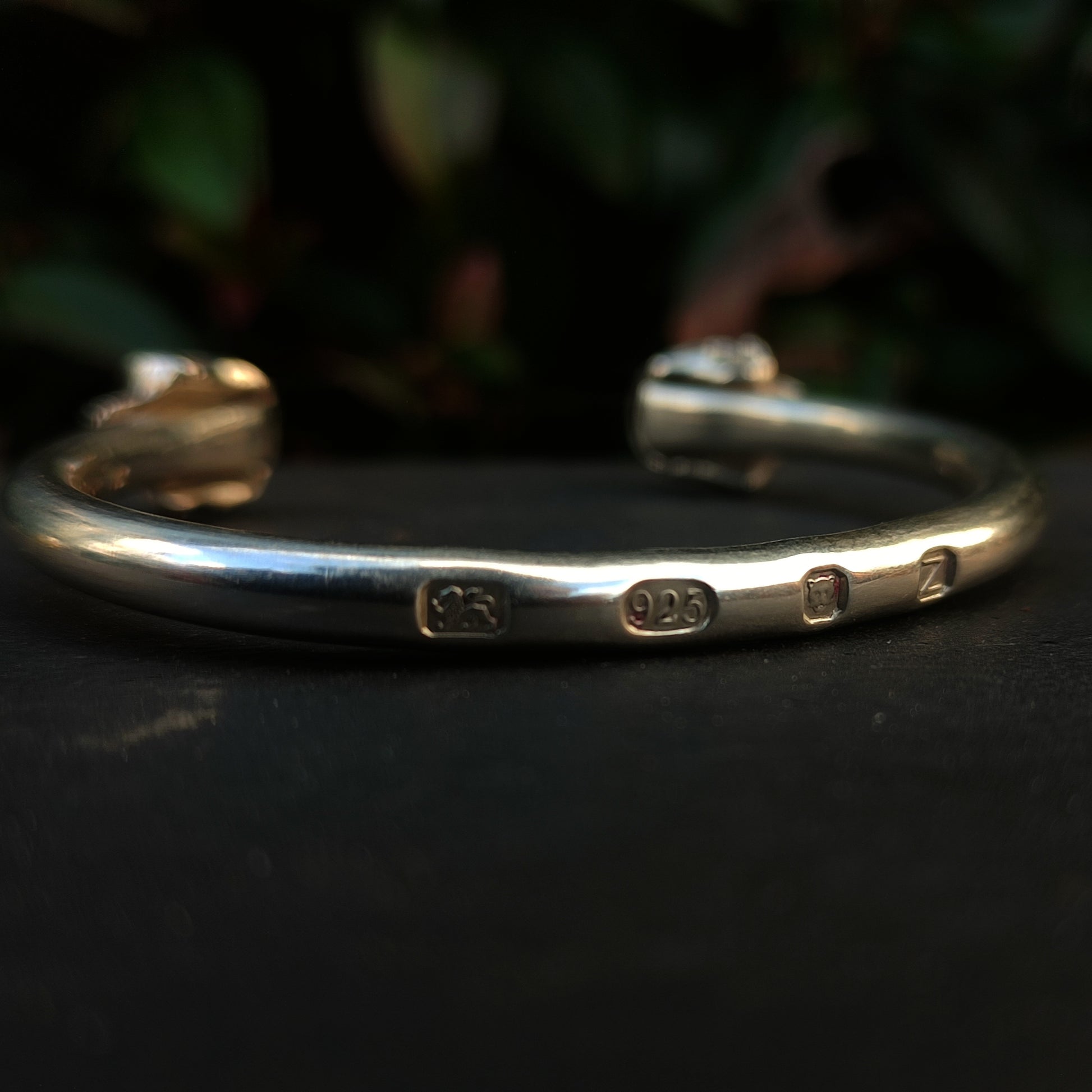 A close-up of the Yellow Sapphire Skull Cuff by silverhollowjewellery, crafted from recycled sterling silver with stamped hallmarks, set against a dark surface and blurred foliage background.