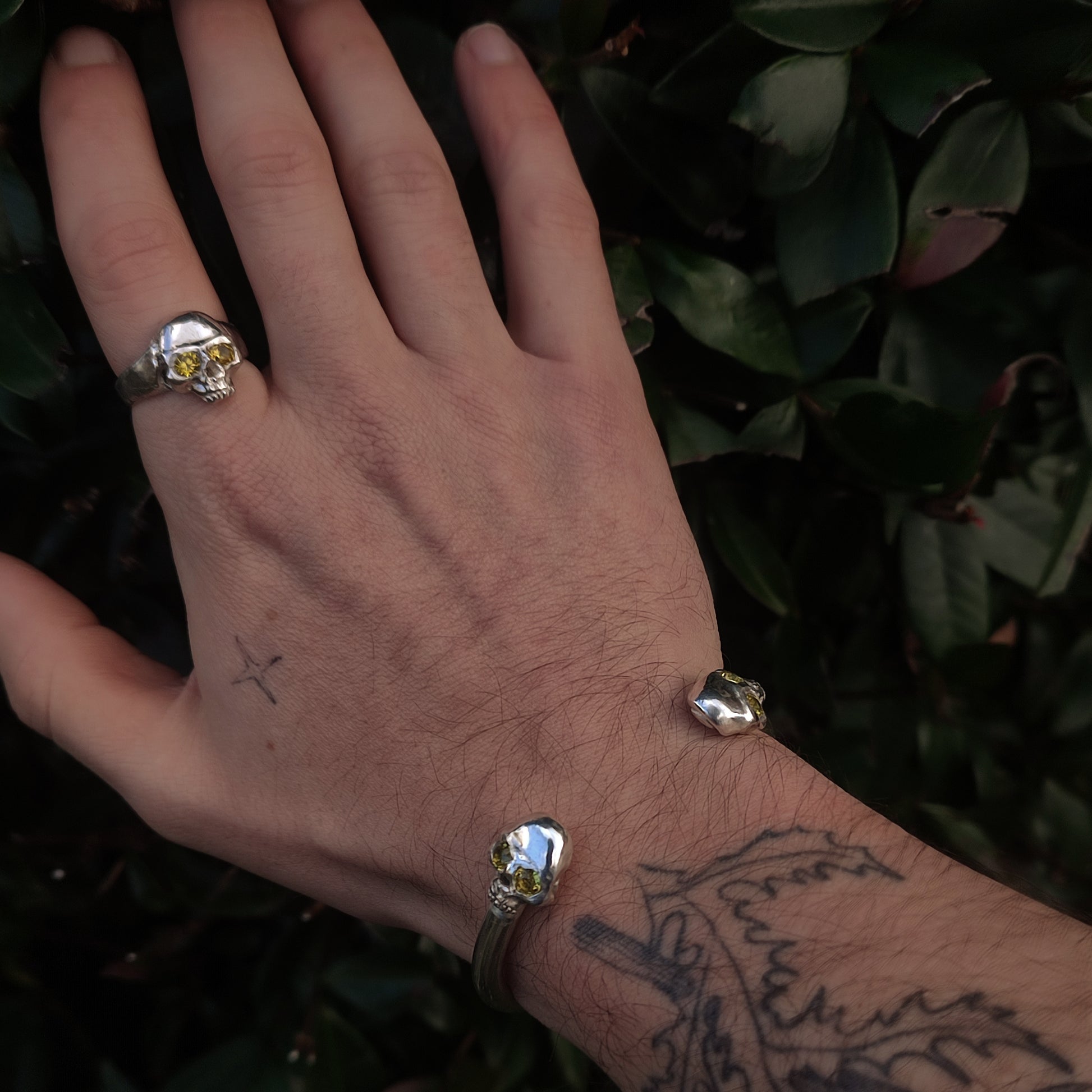 A hand with small cross and wrist tattoos wears the silverhollowjewellery Yellow Sapphire Skull Cuff, set against lush green leaves.