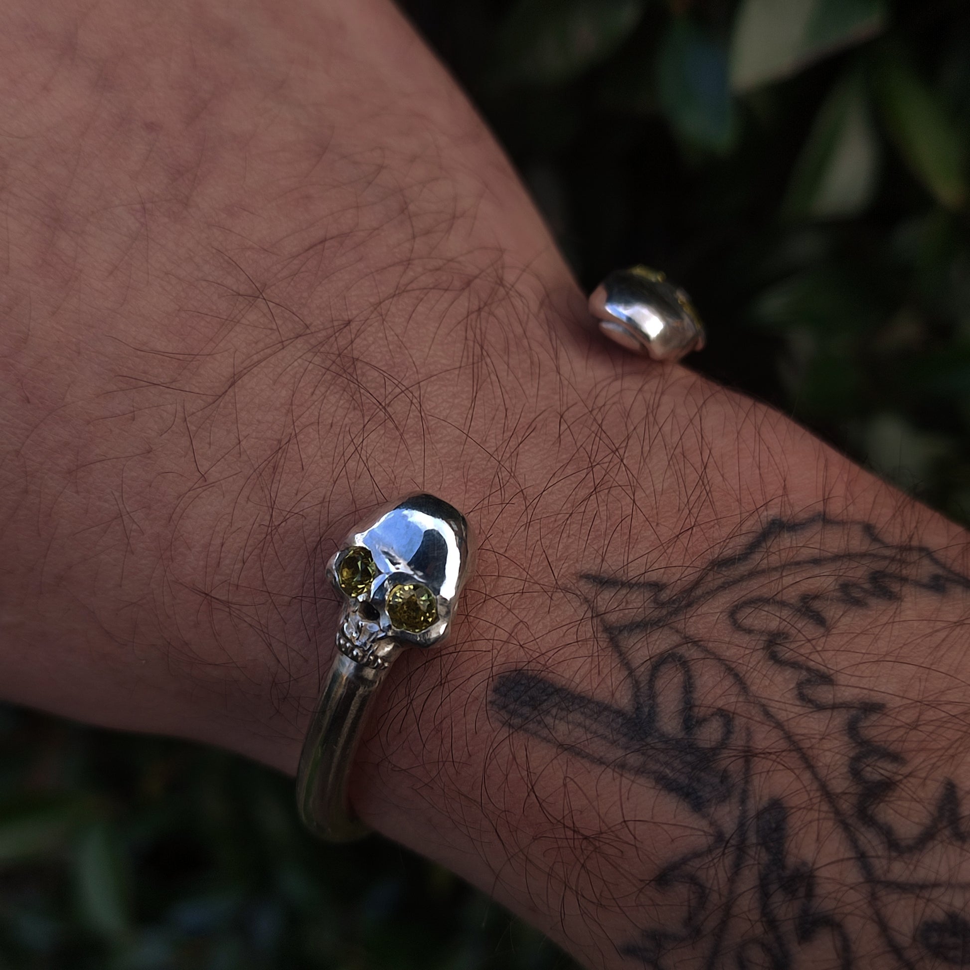 The Yellow Sapphire Skull Cuff by silverhollowjewellery, featuring lab grown yellow sapphires as skull eyes, is showcased on a tattooed wrist.