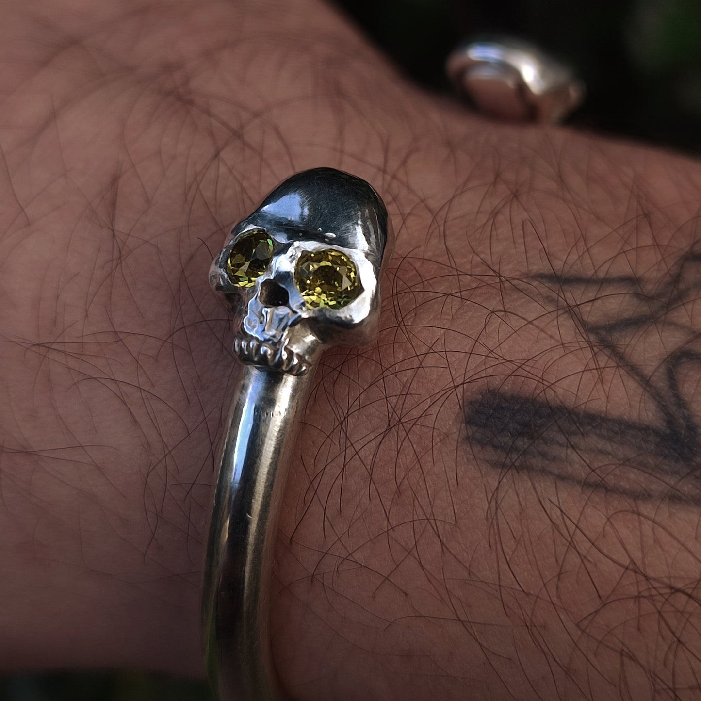 The Yellow Sapphire Skull Cuff by silverhollowjewellery, crafted from recycled sterling silver, features lab-grown yellow sapphire eyes in a skull design and is shown worn on a tattooed wrist.