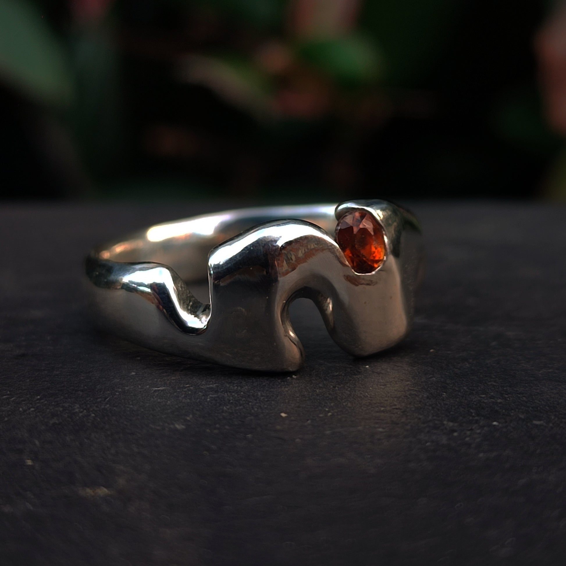 The Sand Cast Sapphire Ring by silverhollowjewellery features a wavy, abstract silver band with an orange oval-cut sapphire, crafted using the traditional sand casting method.