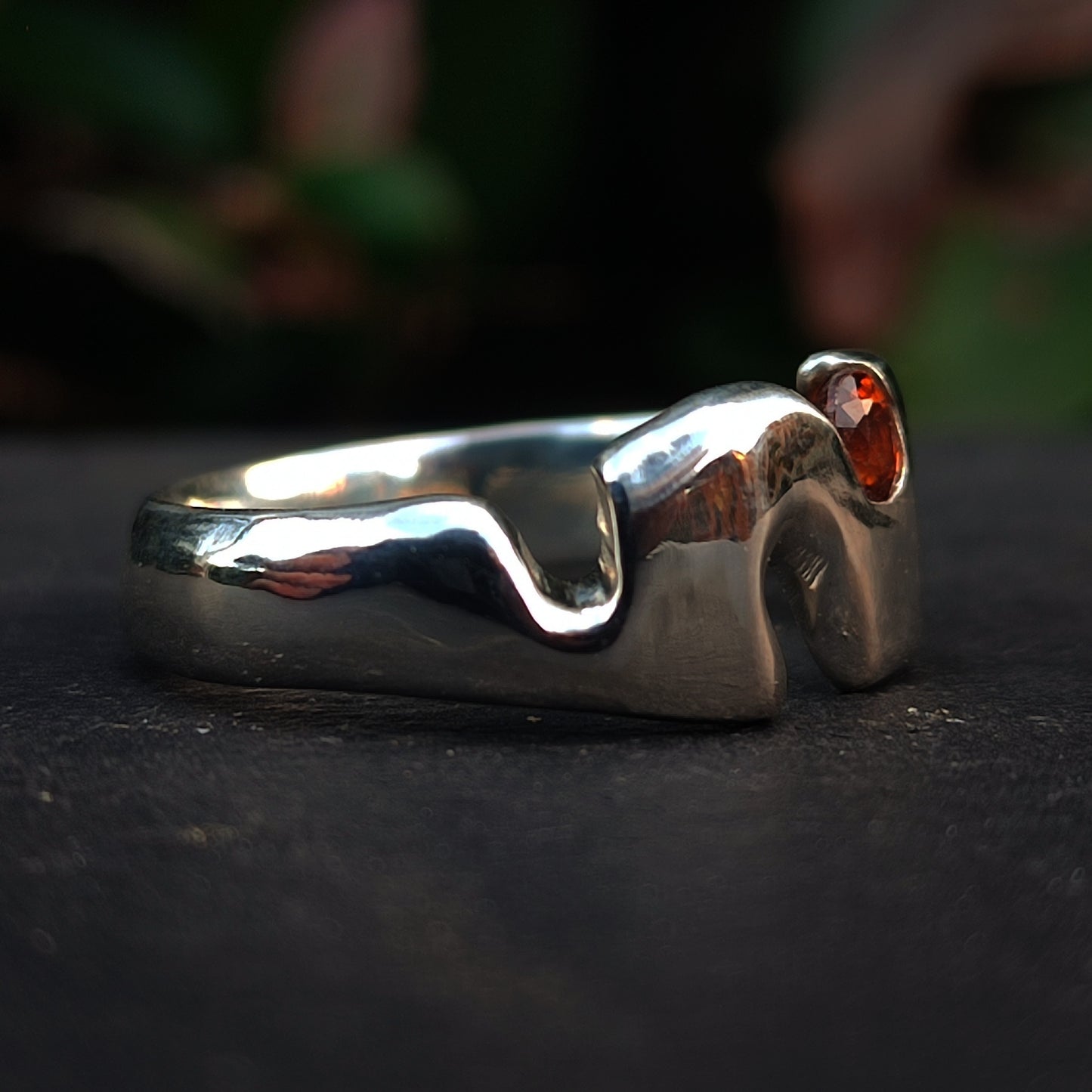 The Sand Cast Sapphire Ring by silverhollowjewellery features an abstract wavy design in recycled sterling silver, set with a small blue sapphire. Shown against a dark blurred background.