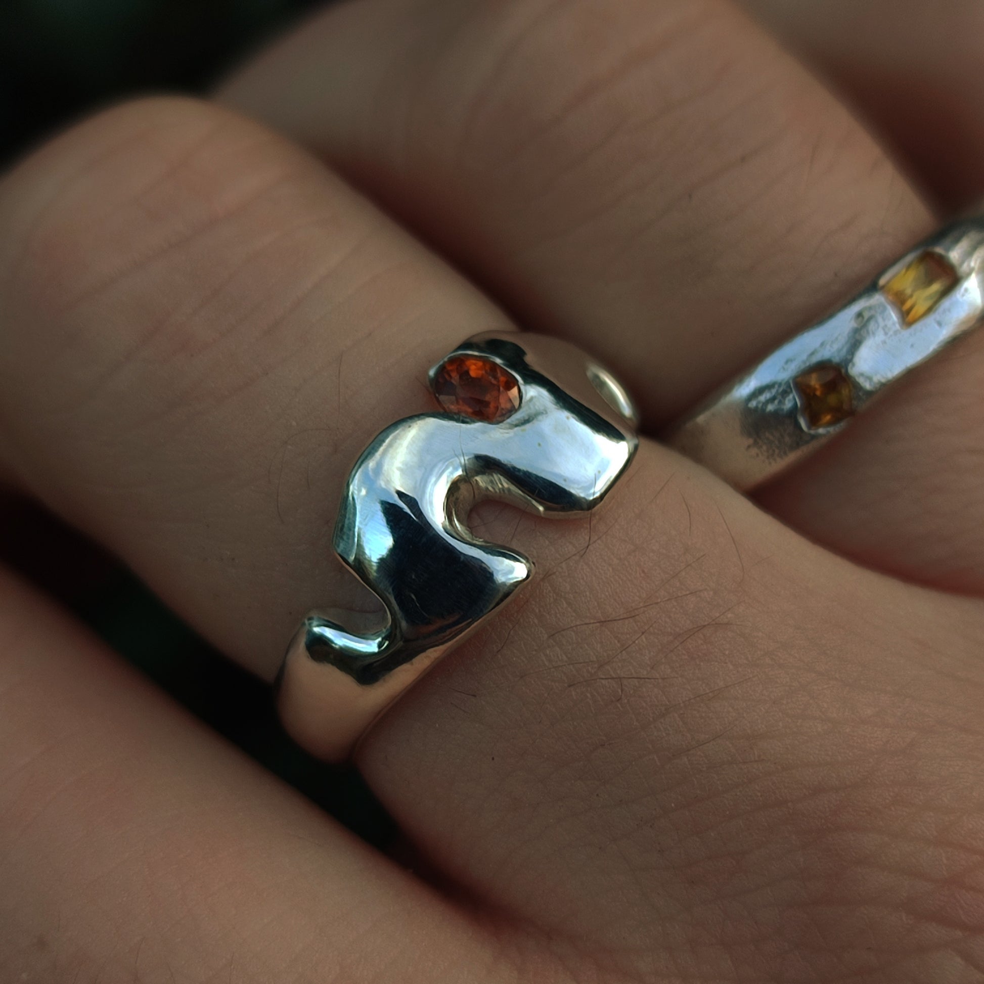 A close-up of two silver rings: the Sand Cast Sapphire Ring by silverhollowjewellery, crafted from recycled sterling silver with an orange sapphire in a stylized animal shape, and another ring featuring small amber-colored stones.
