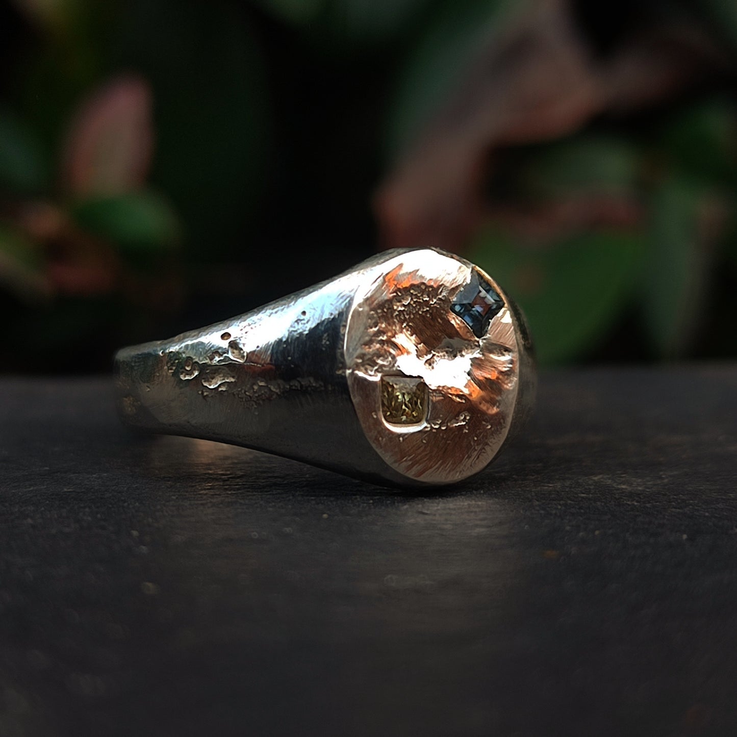 The Sand Cast Sapphire Ring by silverhollowjewellery, featuring a textured, irregular surface crafted with the sand casting method, is showcased on a dark background with blurred greenery.