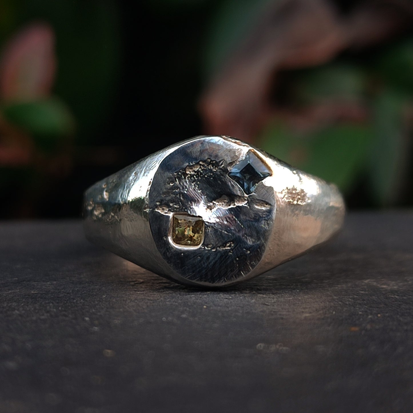 The Sand Cast Sapphire Ring by silverhollowjewellery is a silver ring with a textured circular face, featuring a square yellow stone and a smaller square blue sapphire, crafted using the sand casting method.