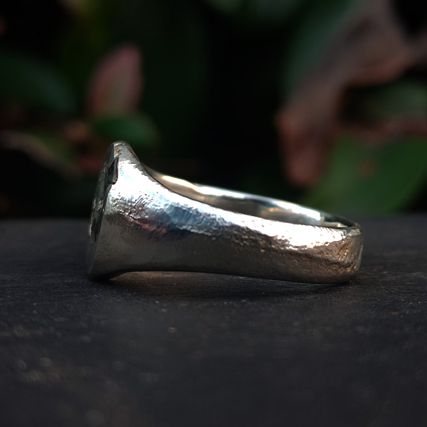 A close-up of the Sand Cast Sapphire Ring by silverhollowjewellery, featuring recycled sterling silver with a textured matte finish, shown resting on a dark surface and blurred greenery in the background.