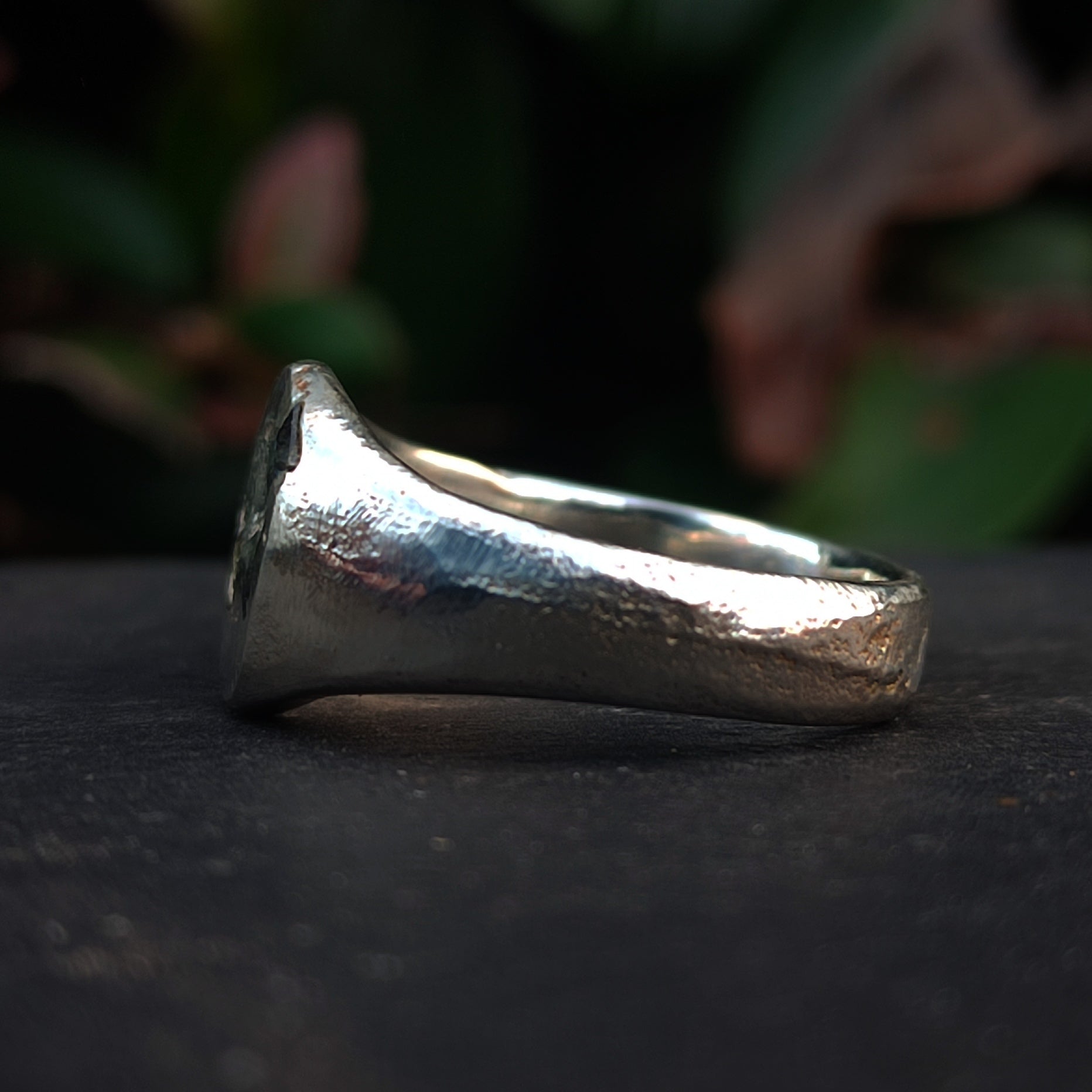 A close-up of the Sand Cast Sapphire Ring by silverhollowjewellery, featuring recycled sterling silver with a textured matte finish, shown resting on a dark surface and blurred greenery in the background.