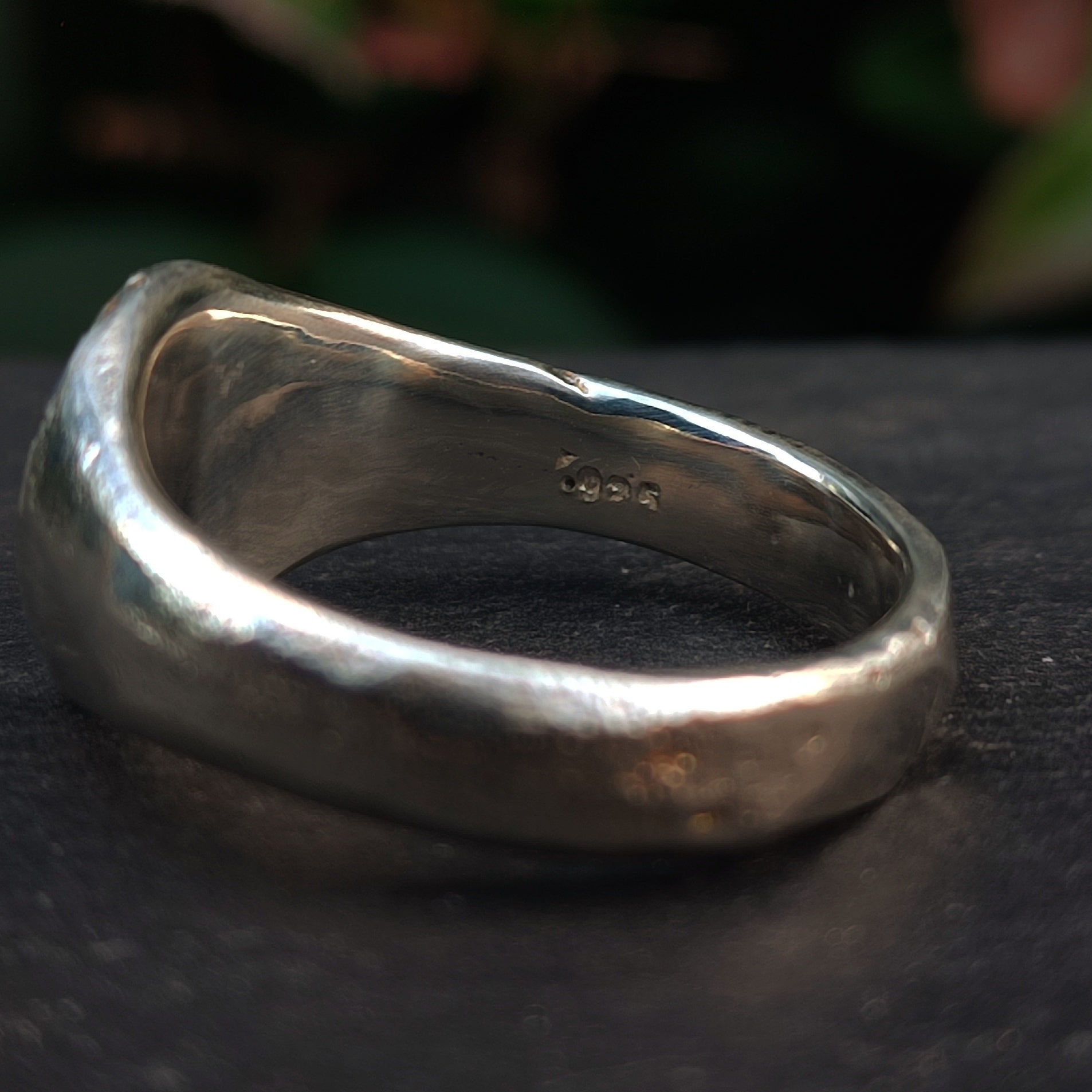 Close-up of the Sand Cast Sapphire Ring by silverhollowjewellery, featuring a smooth, irregular form handcrafted from recycled sterling silver and stamped "925" on the inner band, set on a dark surface with blurred green foliage behind.