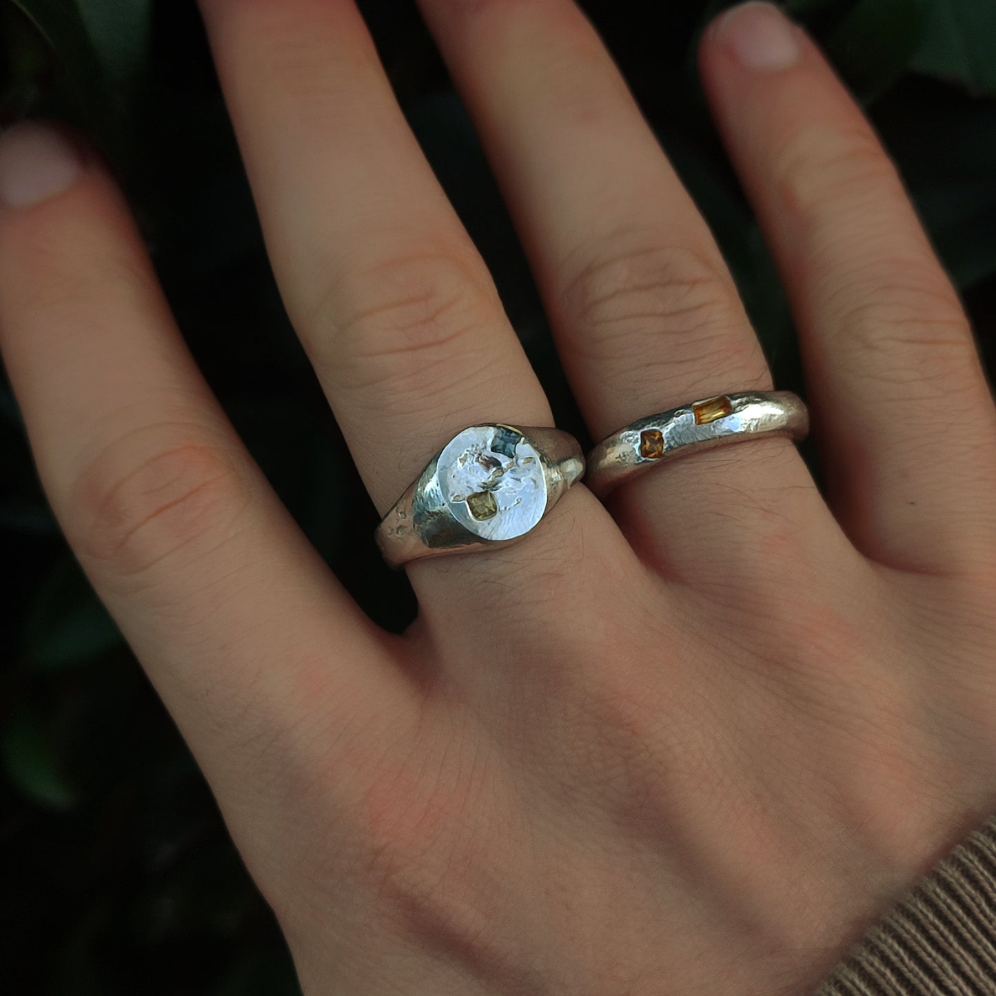 A hand wearing the silverhollowjewellery Sand Cast Sapphire Ring, crafted from recycled sterling silver with embedded sapphires, alongside another ring, set against a leafy background.
