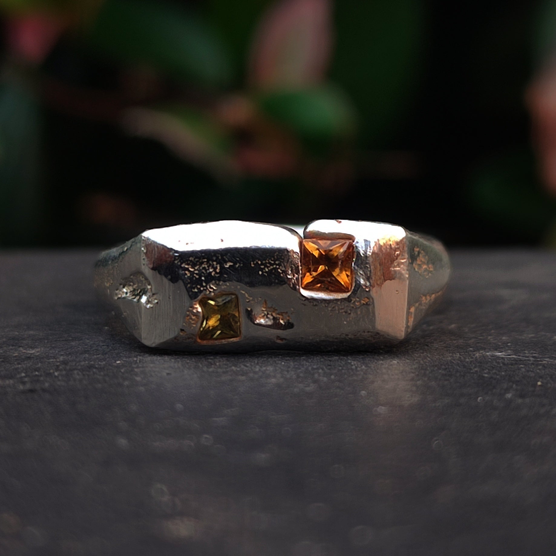 The Sand Cast Sapphire Ring by silverhollowjewellery is a silver ring with a textured band, crafted via sand casting, featuring two square gemstones—one orange and one yellow—set on top.