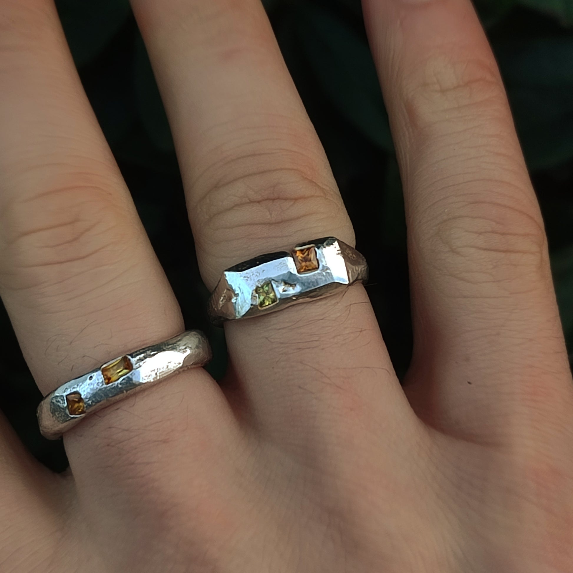 A hand wears two Sand Cast Sapphire Rings by silverhollowjewellery, each featuring a small rectangular yellow princess cut sapphire set in recycled sterling silver, displayed against a dark blurred background.