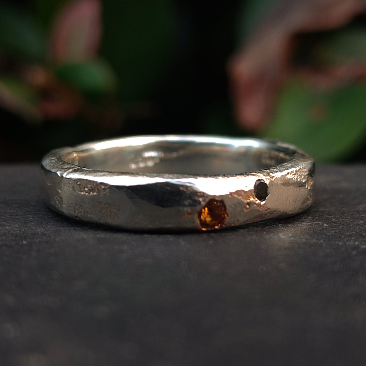 The Sand Cast Sapphire Ring by silverhollowjewellery features a textured silver band crafted with sand casting, set with a small orange sapphire and a distinctive hole. The blurred green background highlights its unique design.