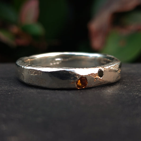 The Sand Cast Sapphire Ring by silverhollowjewellery features a textured silver band crafted with sand casting, set with a small orange sapphire and a distinctive hole. The blurred green background highlights its unique design.