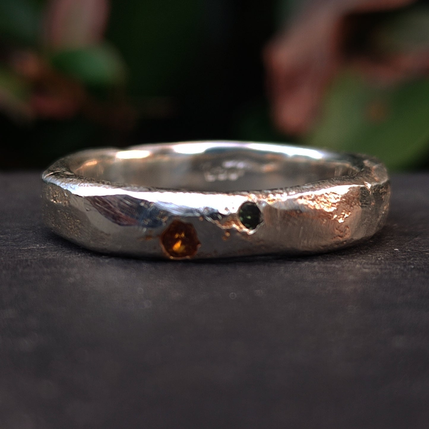 A close-up of the Sand Cast Sapphire Ring by silverhollowjewellery, crafted with textured silver using sand casting, set with a small blue sapphire, displayed on a dark surface with greenery blurred in the background.