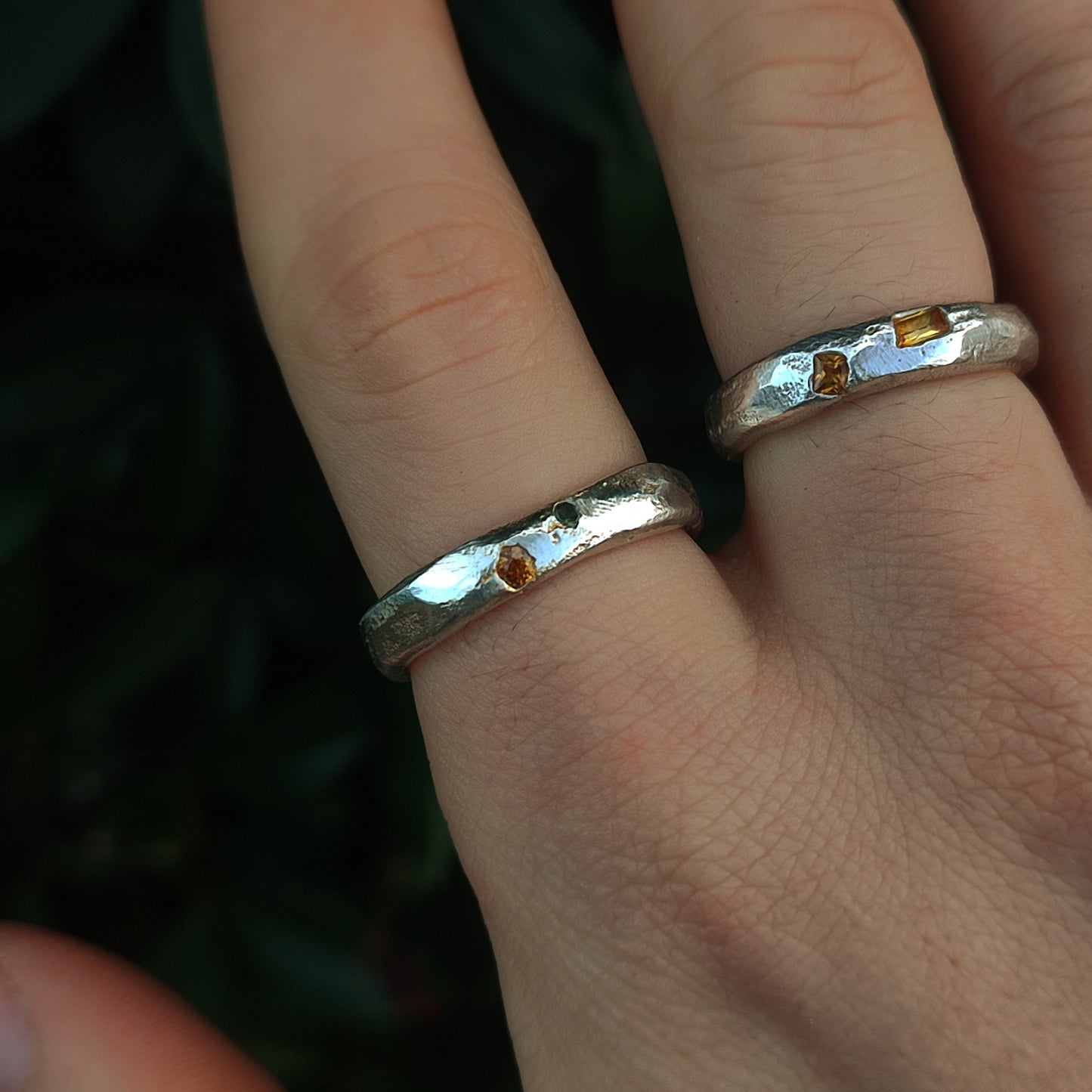 A hand wearing two Sand Cast Sapphire Rings by silverhollowjewellery, crafted from recycled sterling silver and set with small sapphires, is shown against a dark green background.