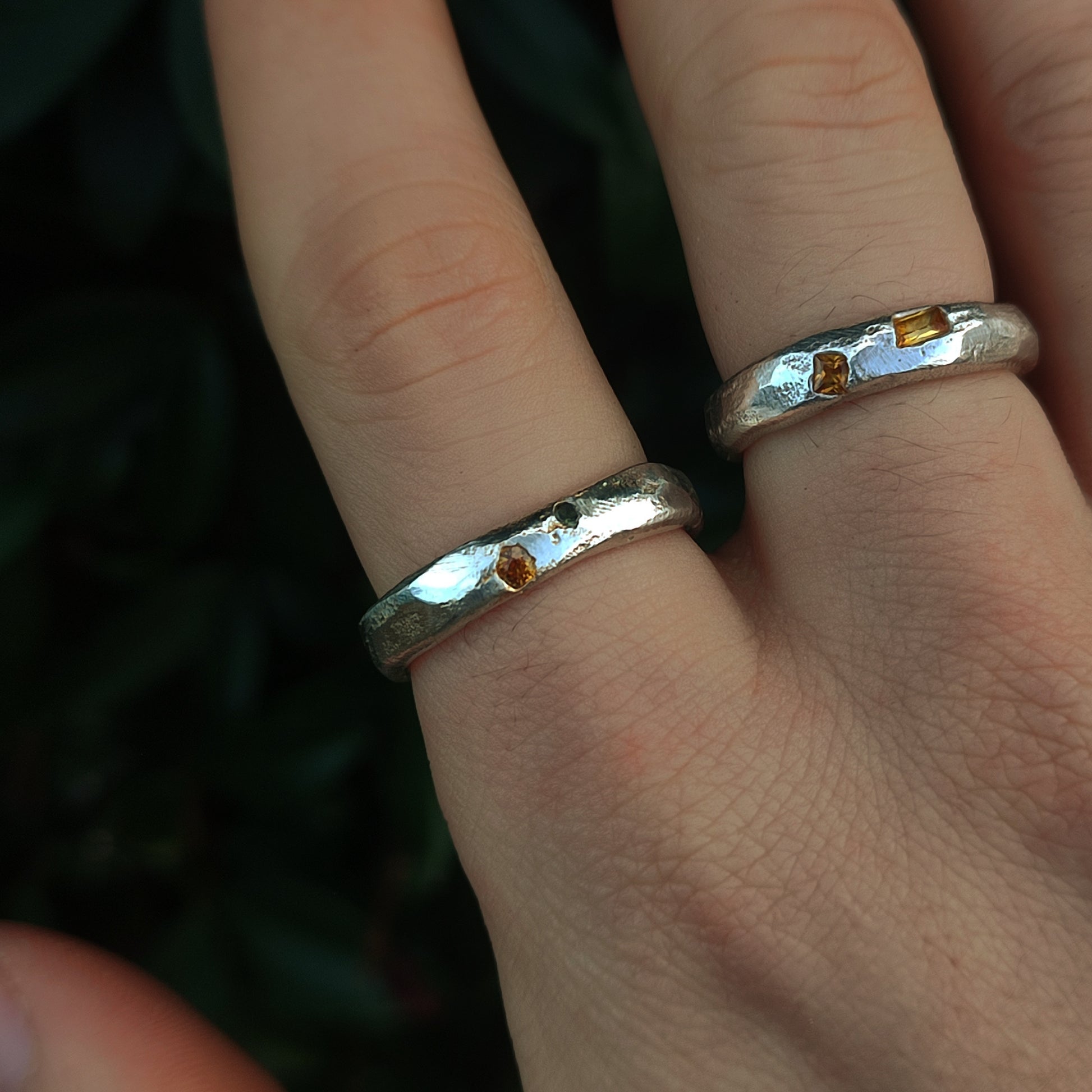 A hand wearing two Sand Cast Sapphire Rings by silverhollowjewellery, crafted from recycled sterling silver and set with small sapphires, is shown against a dark green background.