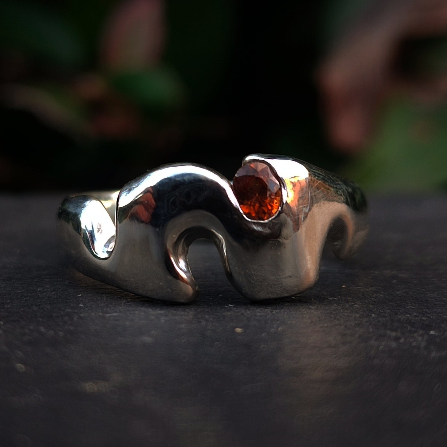 The Sand Cast Sapphire Ring by silverhollowjewellery features a wavy, abstract silver design crafted with traditional sand casting and showcases a small round blue sapphire set near the top.