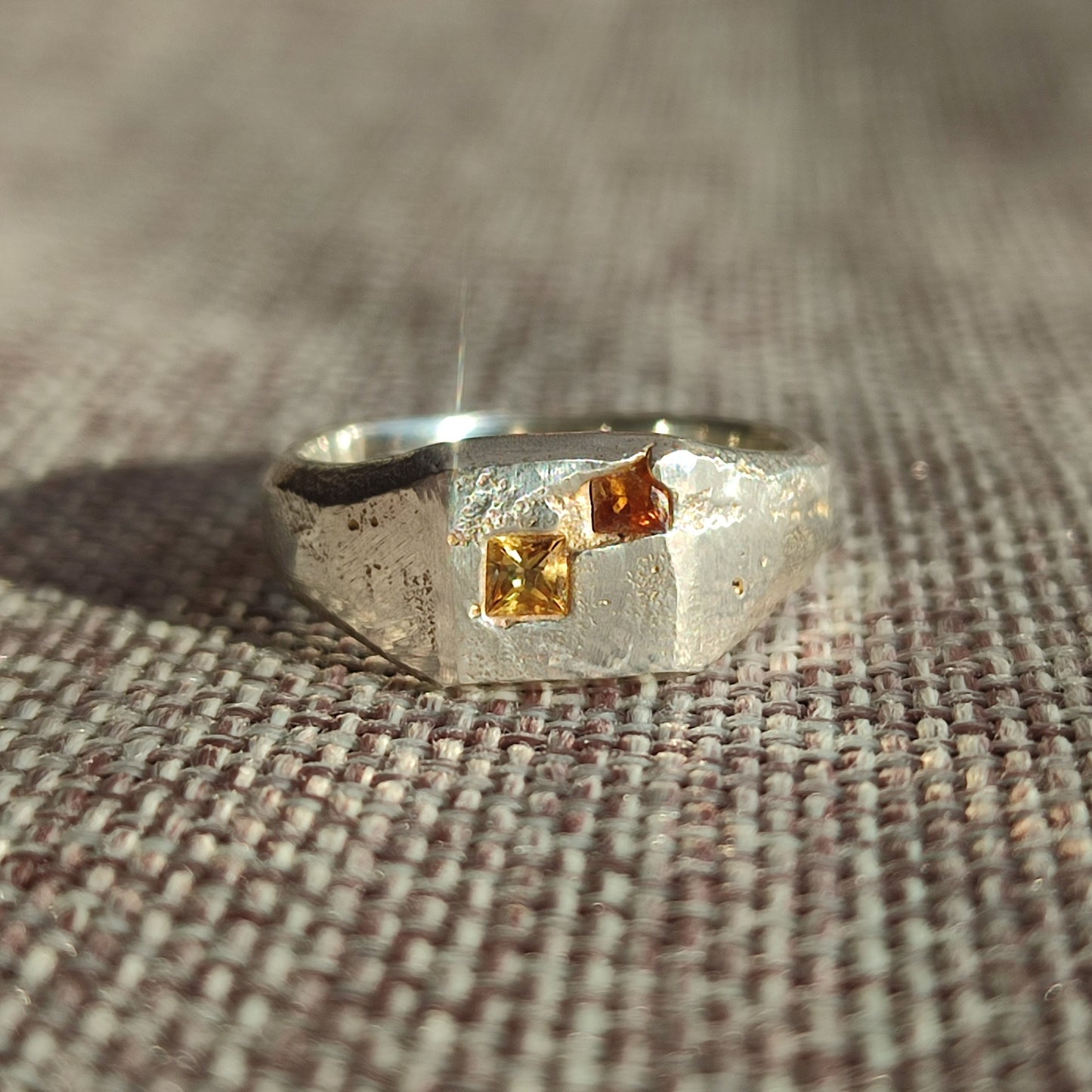 The Sand Cast Sapphire Signet by silverhollowjewellery is a textured recycled sterling silver ring with two square-cut yellow and orange gemstones, shown on woven fabric in natural light.