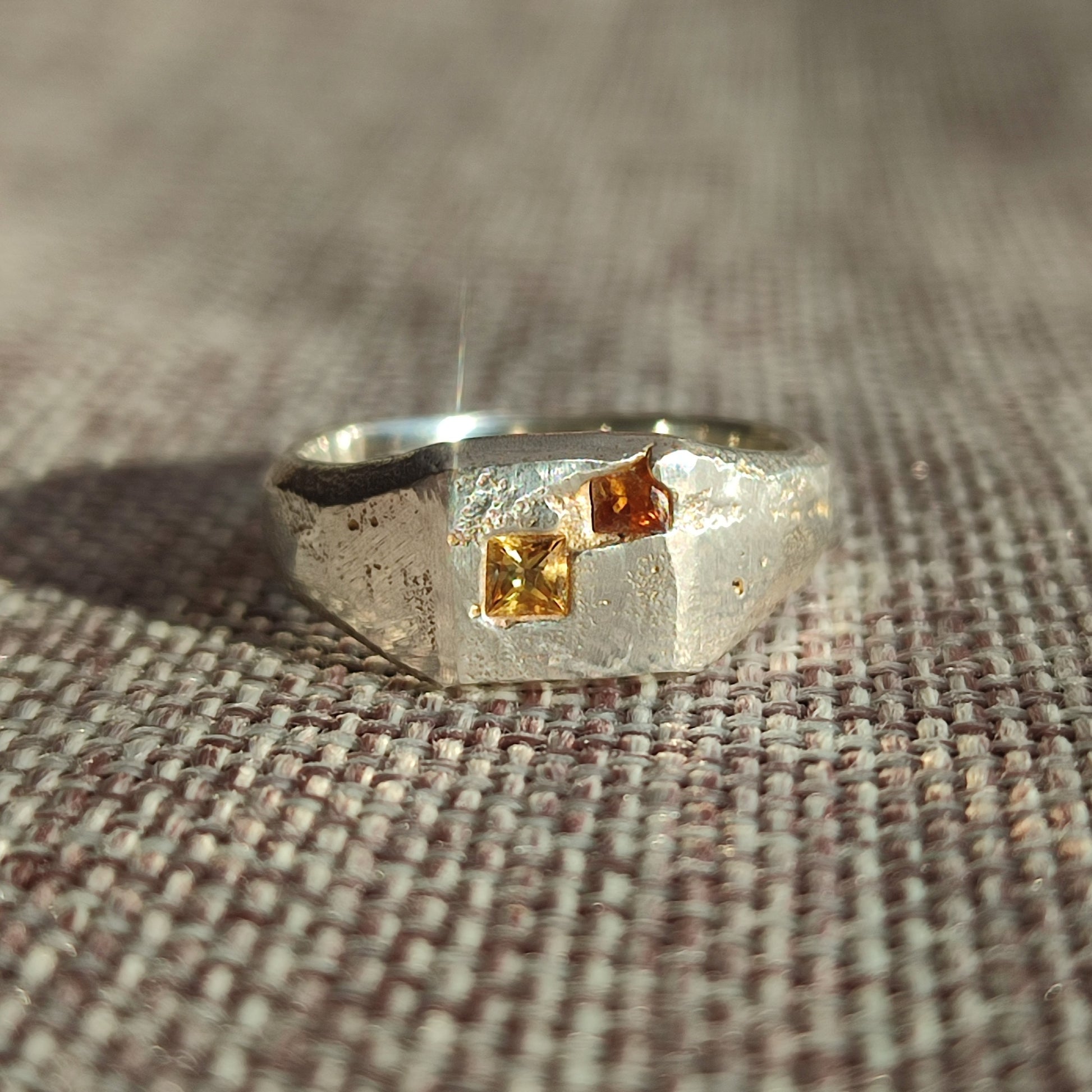 The Sand Cast Sapphire Signet by silverhollowjewellery is a textured recycled sterling silver ring with two square-cut yellow and orange gemstones, shown on woven fabric in natural light.