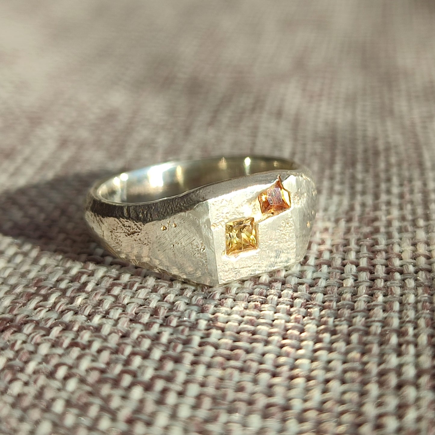 The Sand Cast Sapphire Signet by silverhollowjewellery is a recycled sterling silver ring with a textured finish, featuring two square-cut yellow and pink stones set on a flat surface.