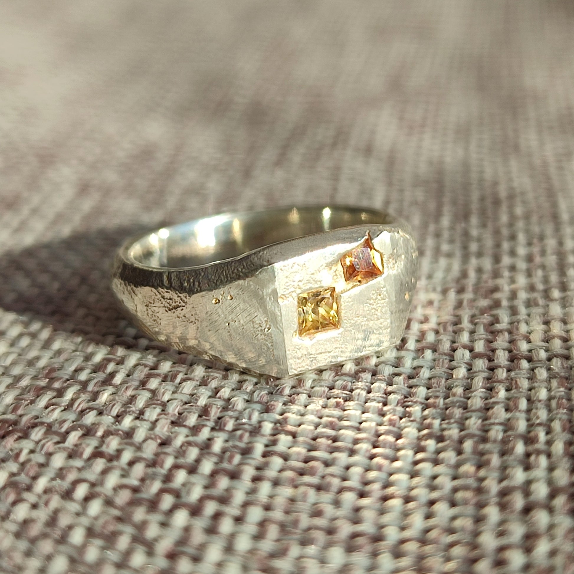 The Sand Cast Sapphire Signet by silverhollowjewellery is a recycled sterling silver ring with a textured finish, featuring two square-cut yellow and pink stones set on a flat surface.