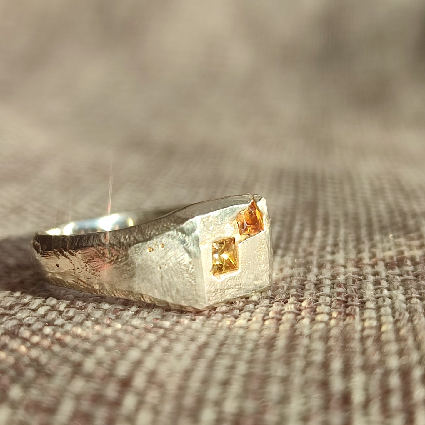 The Sand Cast Sapphire Signet by silverhollowjewellery is a recycled sterling silver ring with a textured band and two small square yellow gemstones set on a flat top, shown resting on woven fabric.
