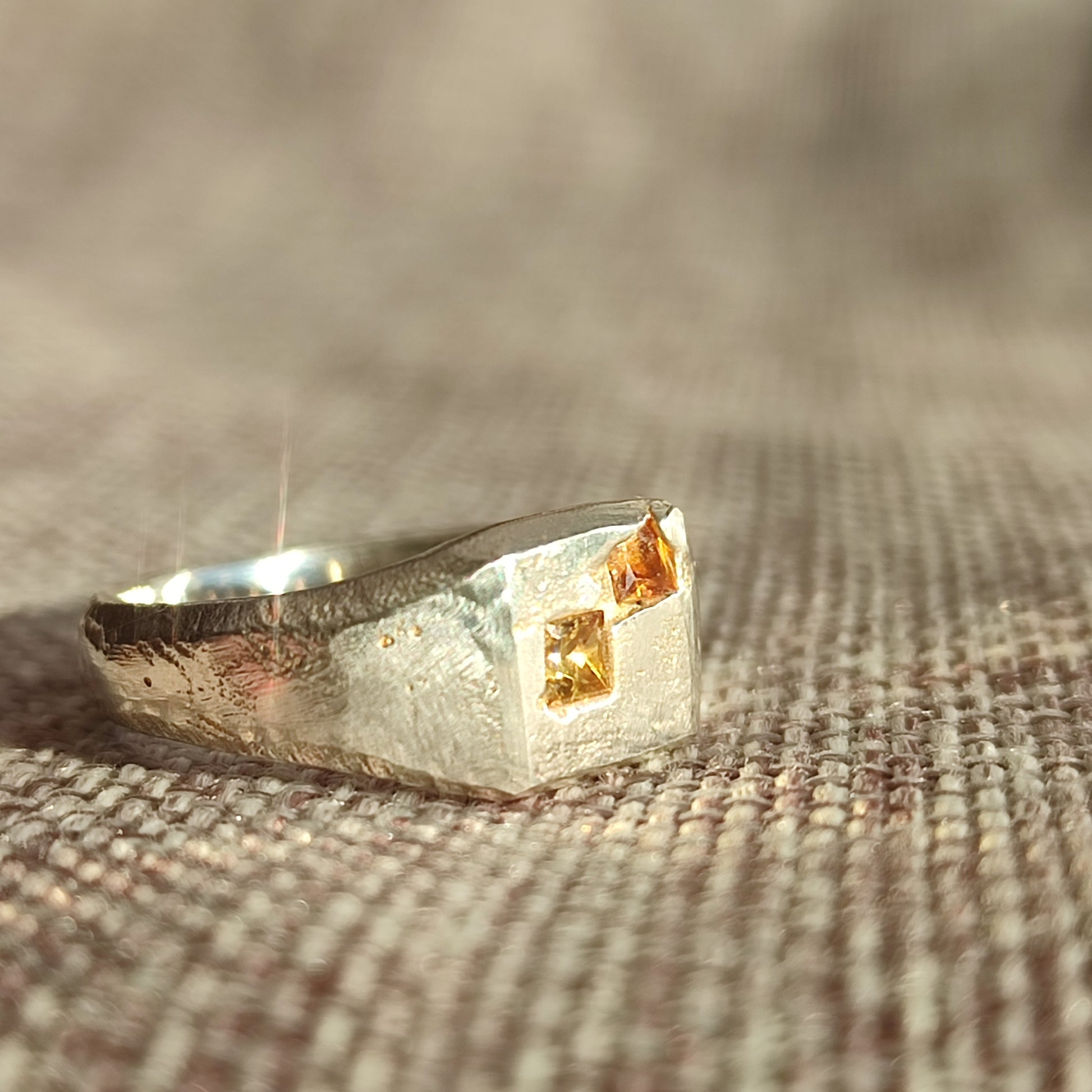 The Sand Cast Sapphire Signet by silverhollowjewellery is a recycled sterling silver ring with a textured band and two small square yellow gemstones set on a flat top, shown resting on woven fabric.