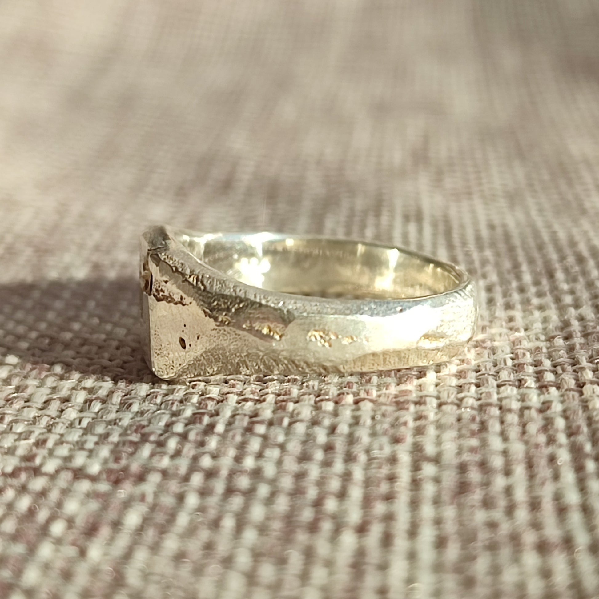 A close-up of the Sand Cast Sapphire Signet by silverhollowjewellery, made from recycled sterling silver with the sand casting method, displayed on woven beige fabric.