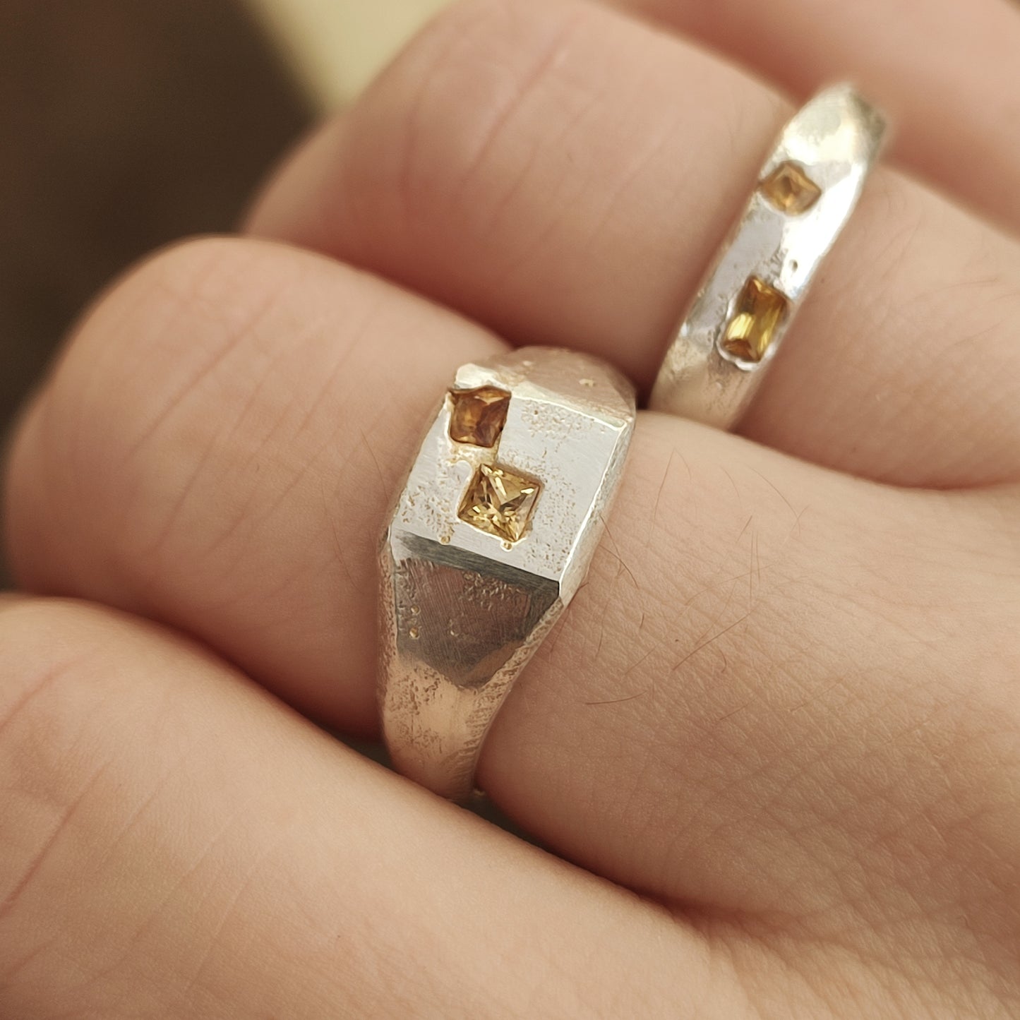 A close-up of two silverhollowjewellery Sand Cast Sapphire Signet rings, featuring yellow princess cut sapphires and crafted from recycled sterling silver, worn on adjacent fingers.