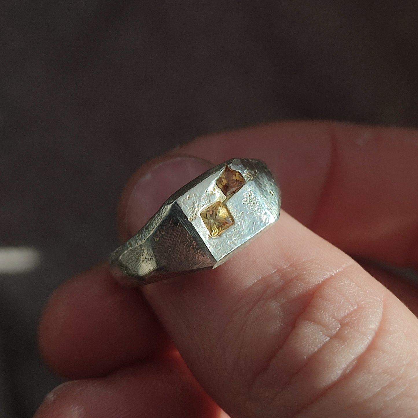 A hand holds the Sand Cast Sapphire Signet by silverhollowjewellery, a geometric recycled sterling silver ring set with two small, square yellow princess cut sapphires on top.